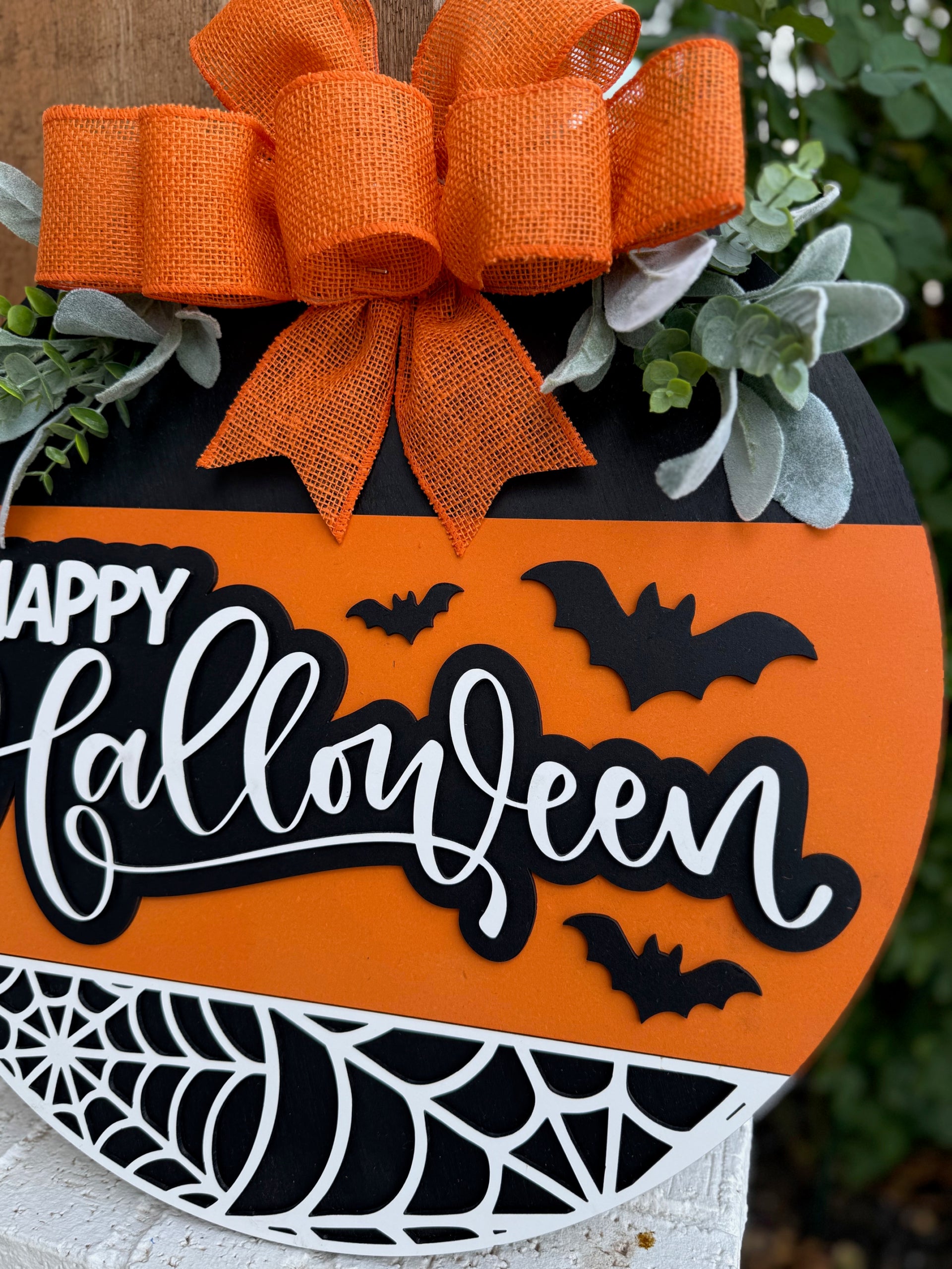 A round Halloween sign with orange, black, and white colors, decorated with black bats, a spiderweb, green leaves, and a large orange burlap bow. The sign reads “Happy Halloween” in white cursive letters.