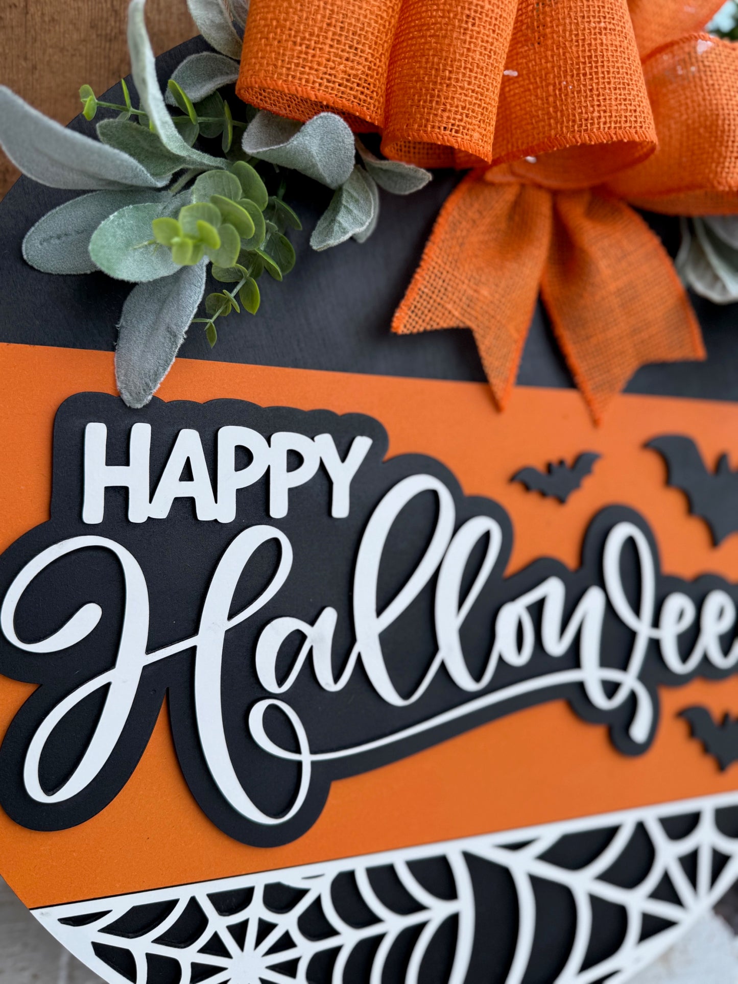 A close-up of a Halloween decoration with Happy Halloween in white script on black and orange background, accented by black bats, a spider web, green leaves, and an orange burlap bow at the top.