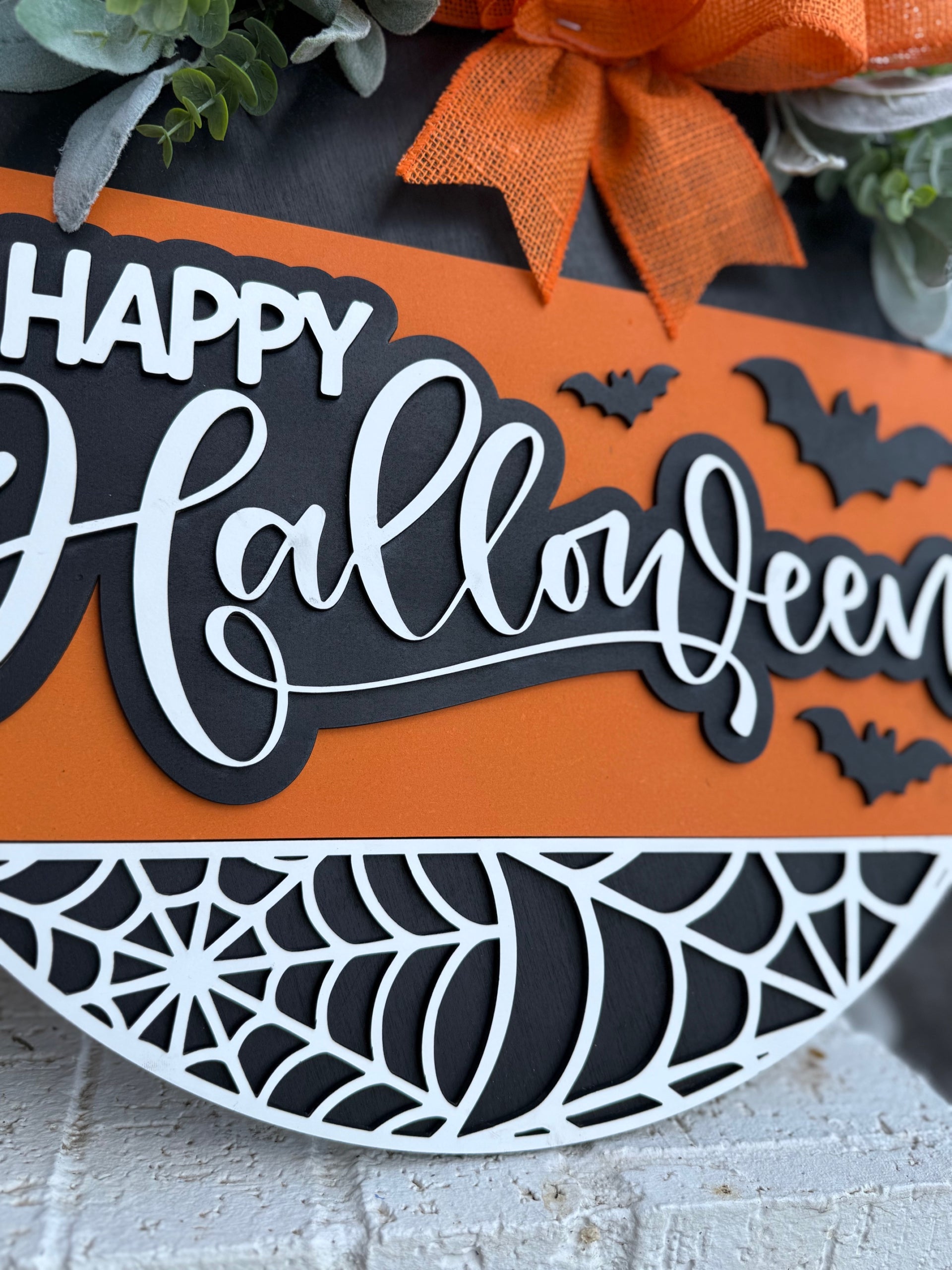 Close-up of a decorative sign reading Happy Halloween in white script on an orange background, with black bats and a white spiderweb design. An orange ribbon and greenery are visible at the top.