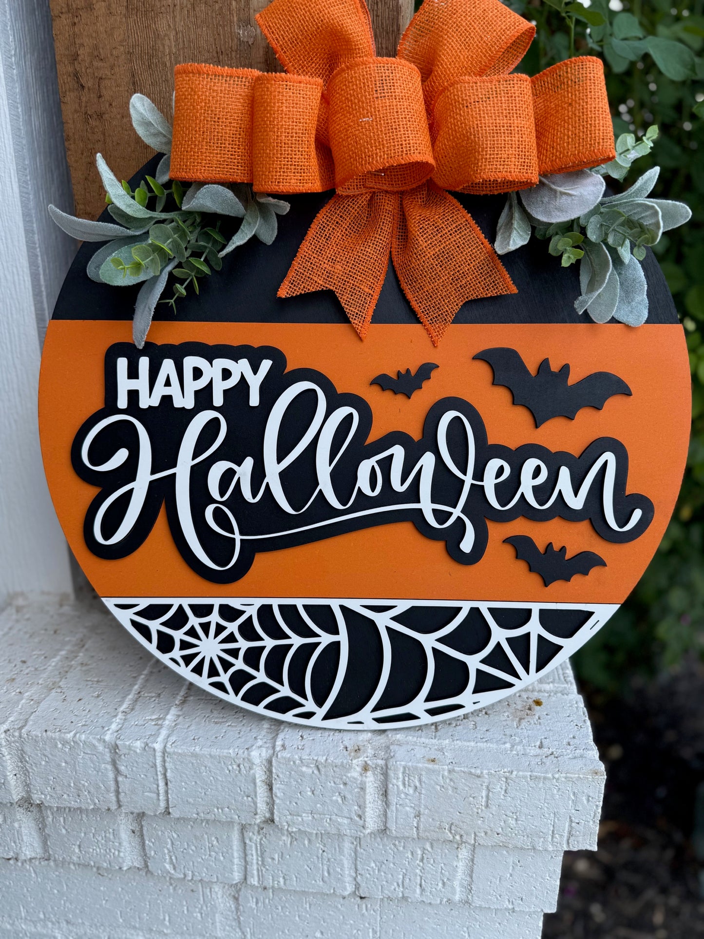 Round Halloween sign with orange and black stripes, decorated with a large orange bow, green leaves, black bats, and a white spiderweb. The words Happy Halloween are written in bold white script.
