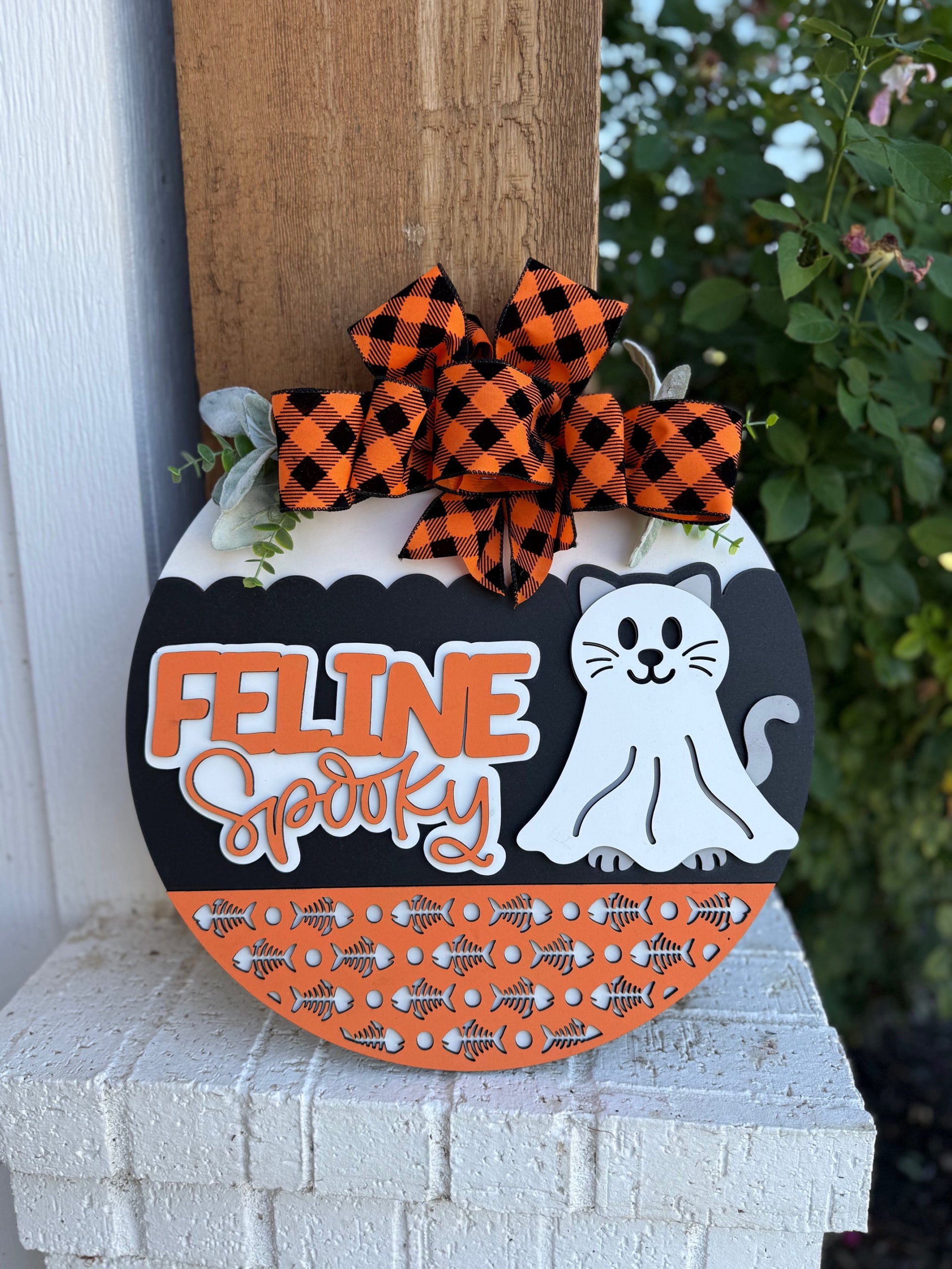 A round Halloween sign decorated with a black-and-white cat, the words “Feline Spooky,” orange-and-black patterns with fishbones, and a large orange plaid bow on top, hanging outdoors by a white wall and green plants.