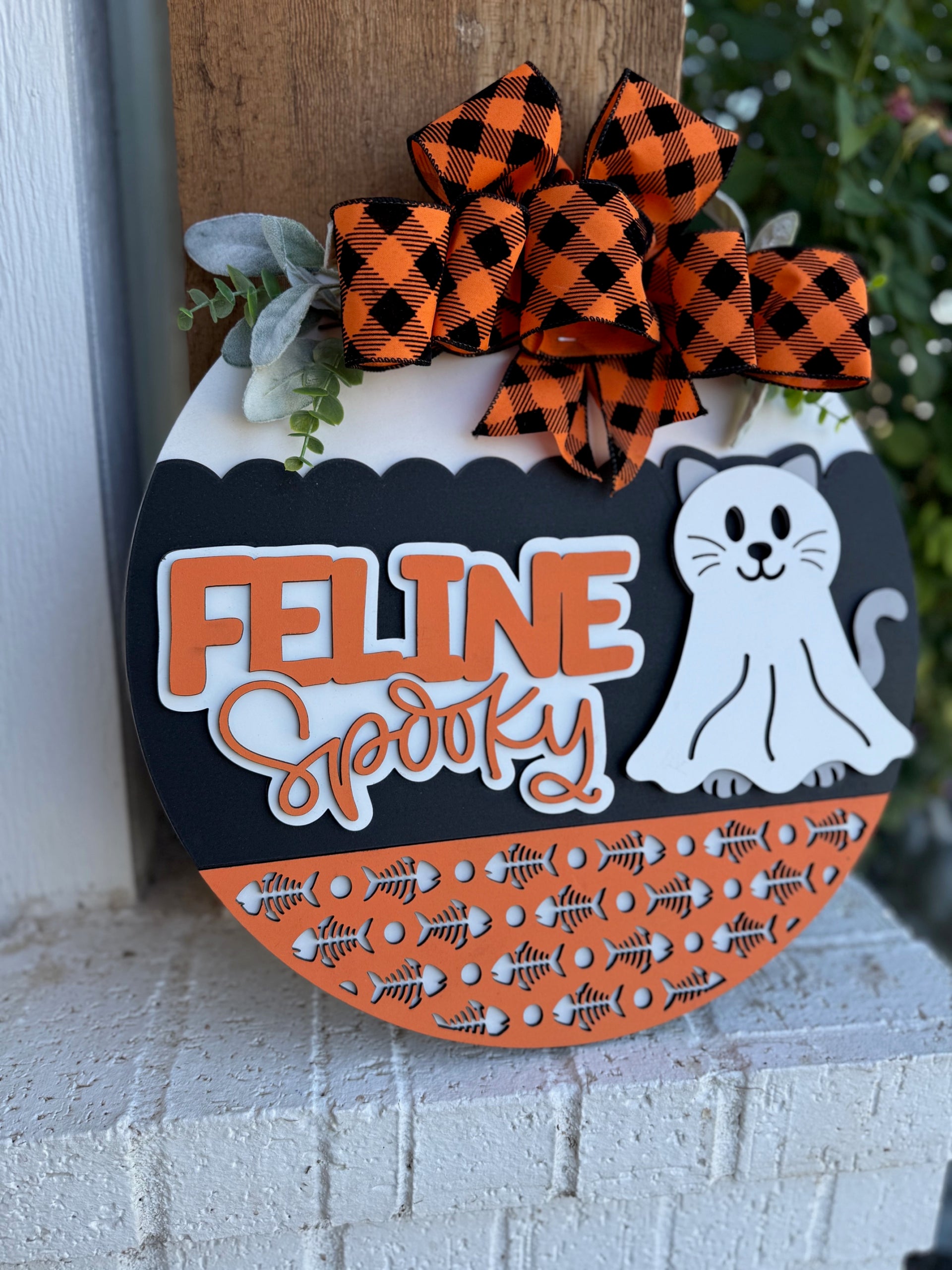 A decorative round sign features a white cat, the words “Feline Spooky,” orange fishbone patterns, green leaves, and a black-and-orange plaid bow. The sign has a Halloween theme and rests on a white brick surface.
