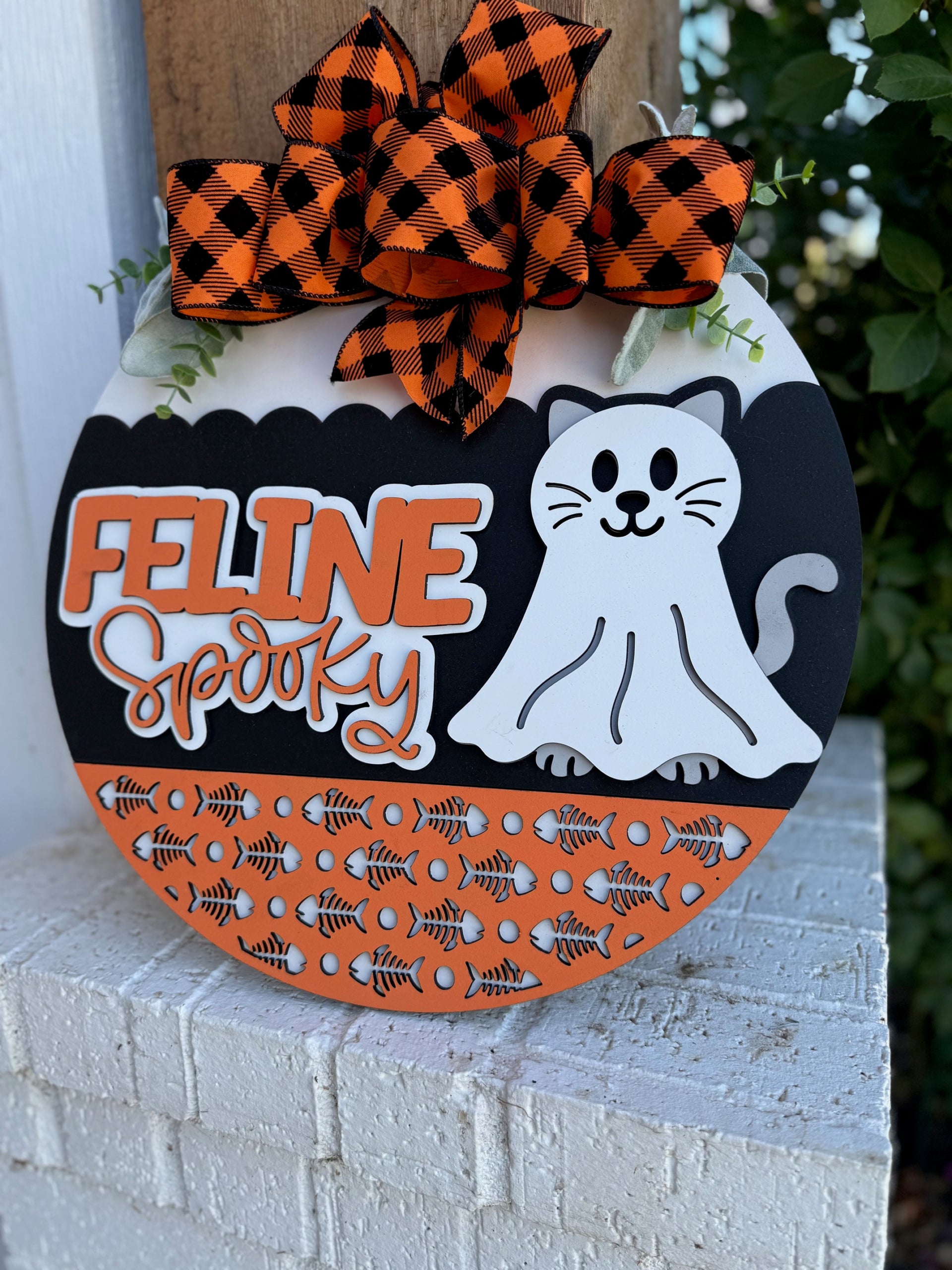 A round Halloween sign with an orange and black plaid bow, the words Feline Spooky, a white cartoon cat, and an orange section with fishbone patterns, set against a brick ledge and greenery.