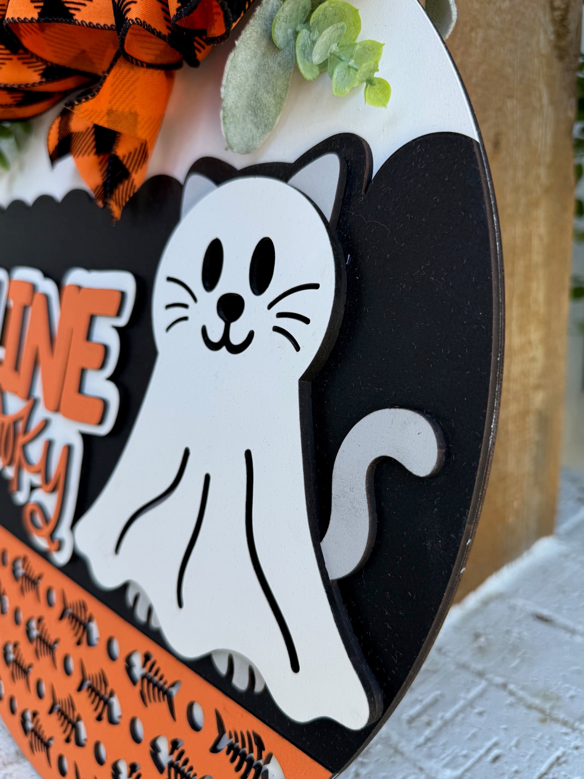 A close-up of a Halloween decoration featuring a cartoon cat dressed as a ghost, with a white sheet, black details, and a tail. The background includes orange and black patterns and a buffalo plaid bow.