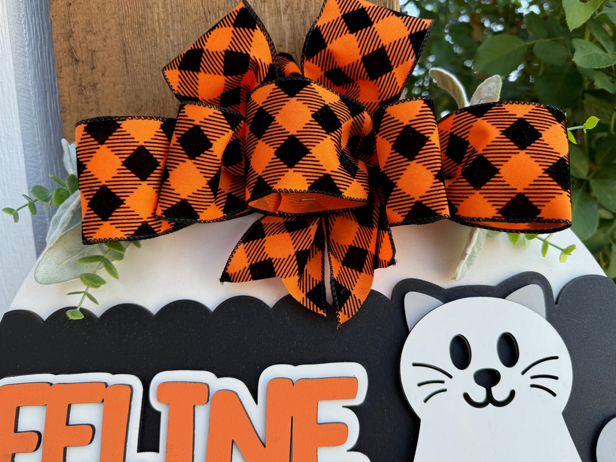 A close-up of an orange and black plaid bow atop a sign featuring a cute white cartoon cat, black scalloped trim, greenery accents, and partially visible bold orange lettering.
