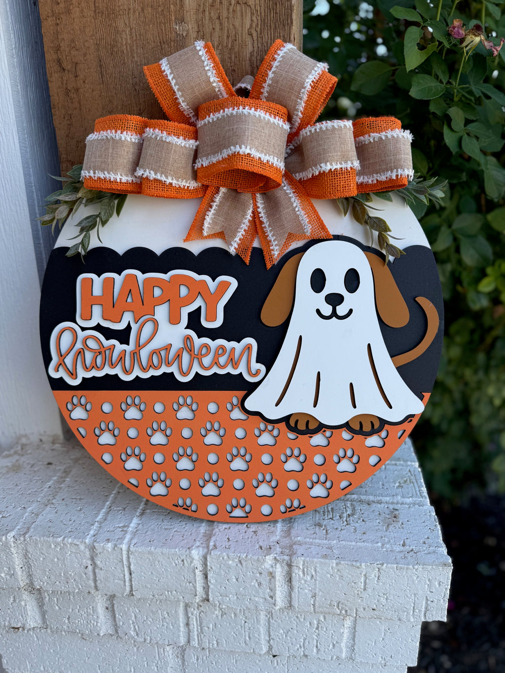 Round Halloween sign decorated with orange and white paw prints, a dog dressed as a ghost, and the text Happy Halloween. An orange and beige bow sits at the top, and greenery is around the edges.