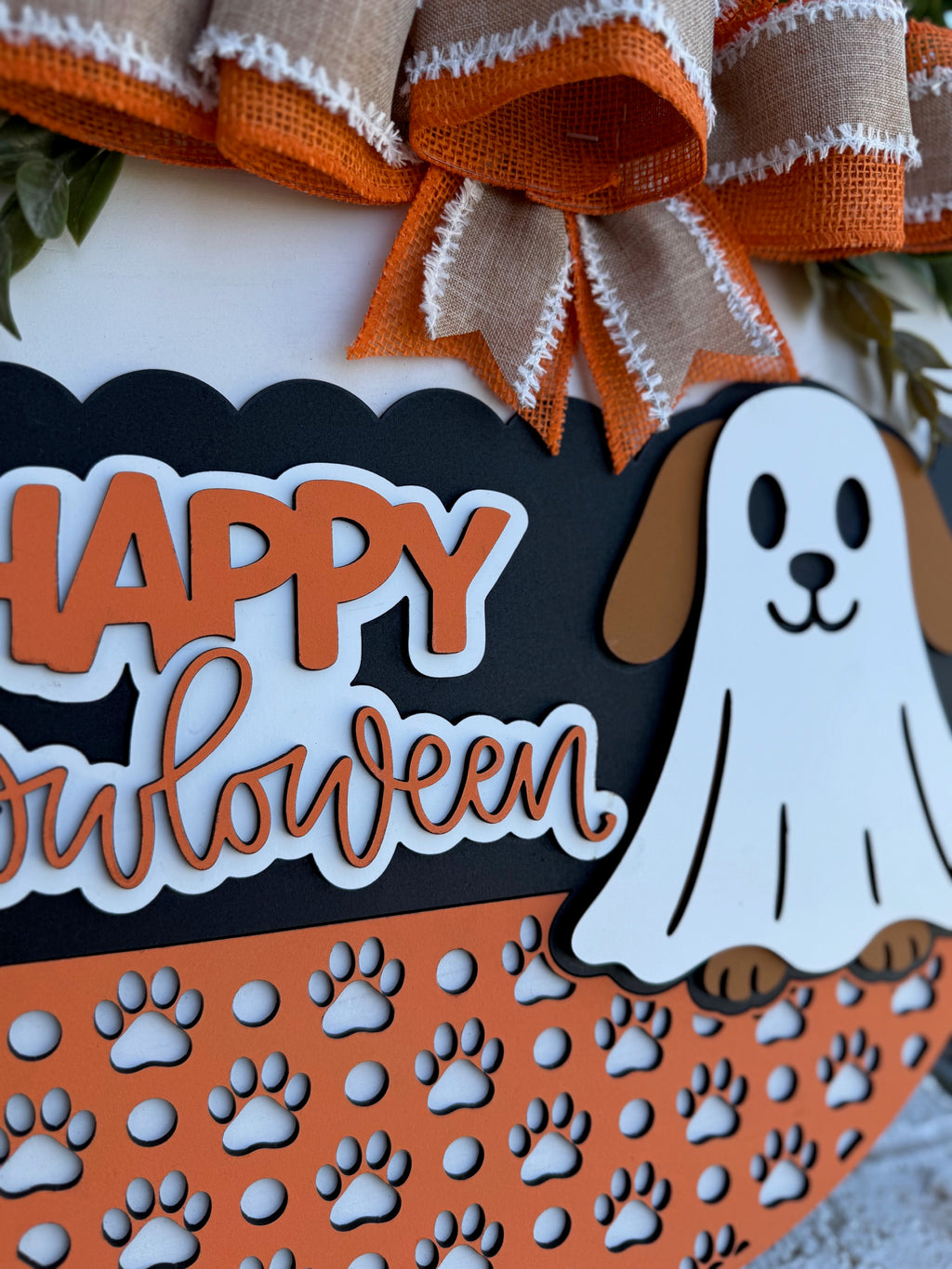 A Halloween decoration features a sign that reads Happy Howloween with a cartoon dog dressed as a ghost. The sign is orange, black, and white with paw prints and burlap bows as accents.