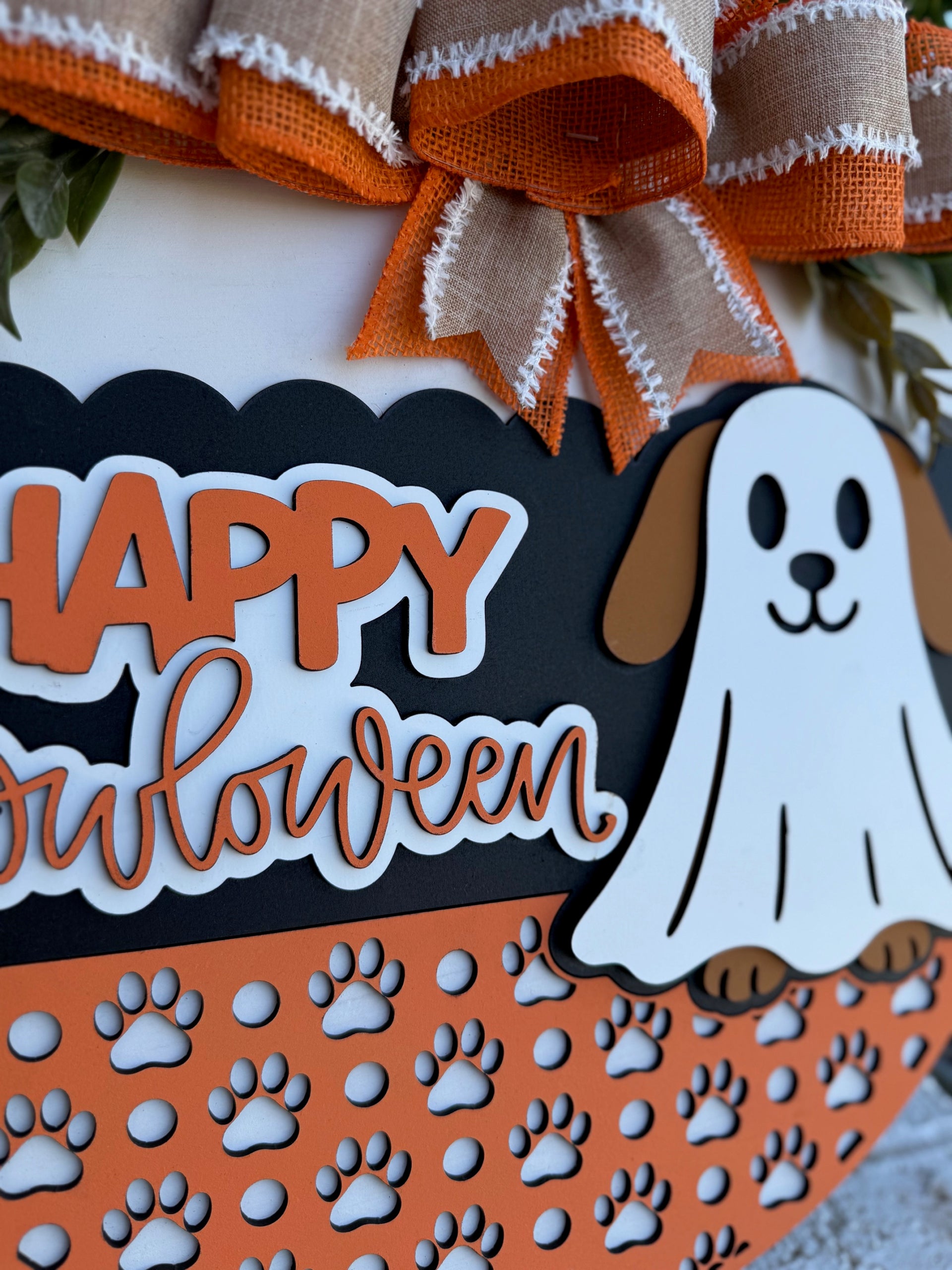 A Halloween decoration features a sign that reads Happy Howloween with a cartoon dog dressed as a ghost. The sign is orange, black, and white with paw prints and burlap bows as accents.