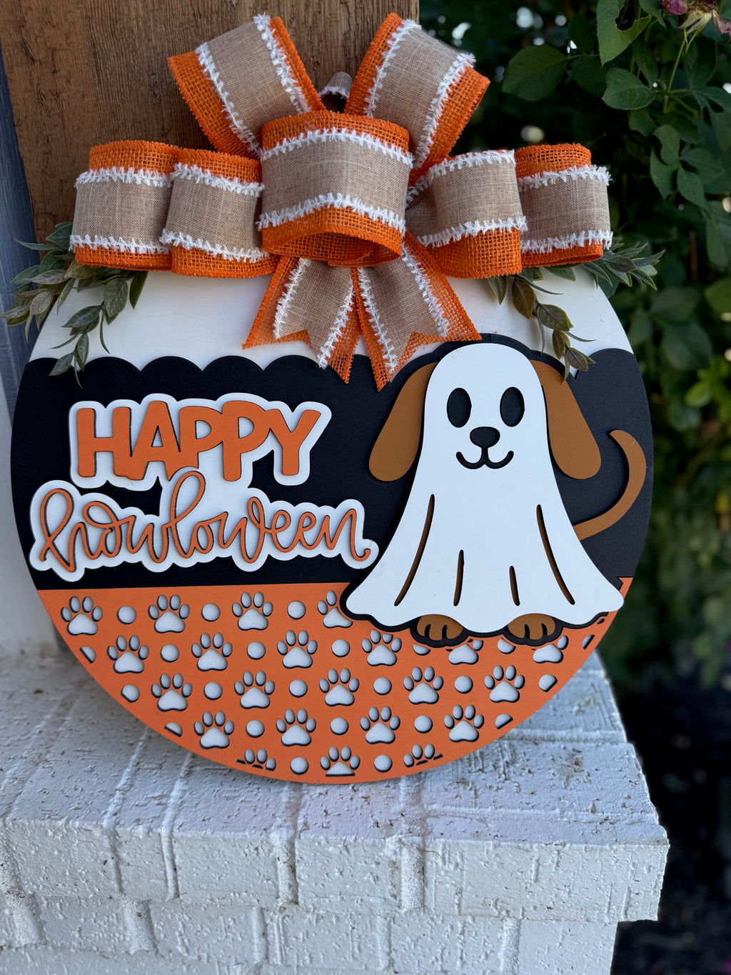 A Halloween-themed round sign with a cartoon dog in a ghost costume, orange bow on top, and the words Happy Howloween above a pattern of white paw prints. The sign rests on a white brick surface with greenery behind it.