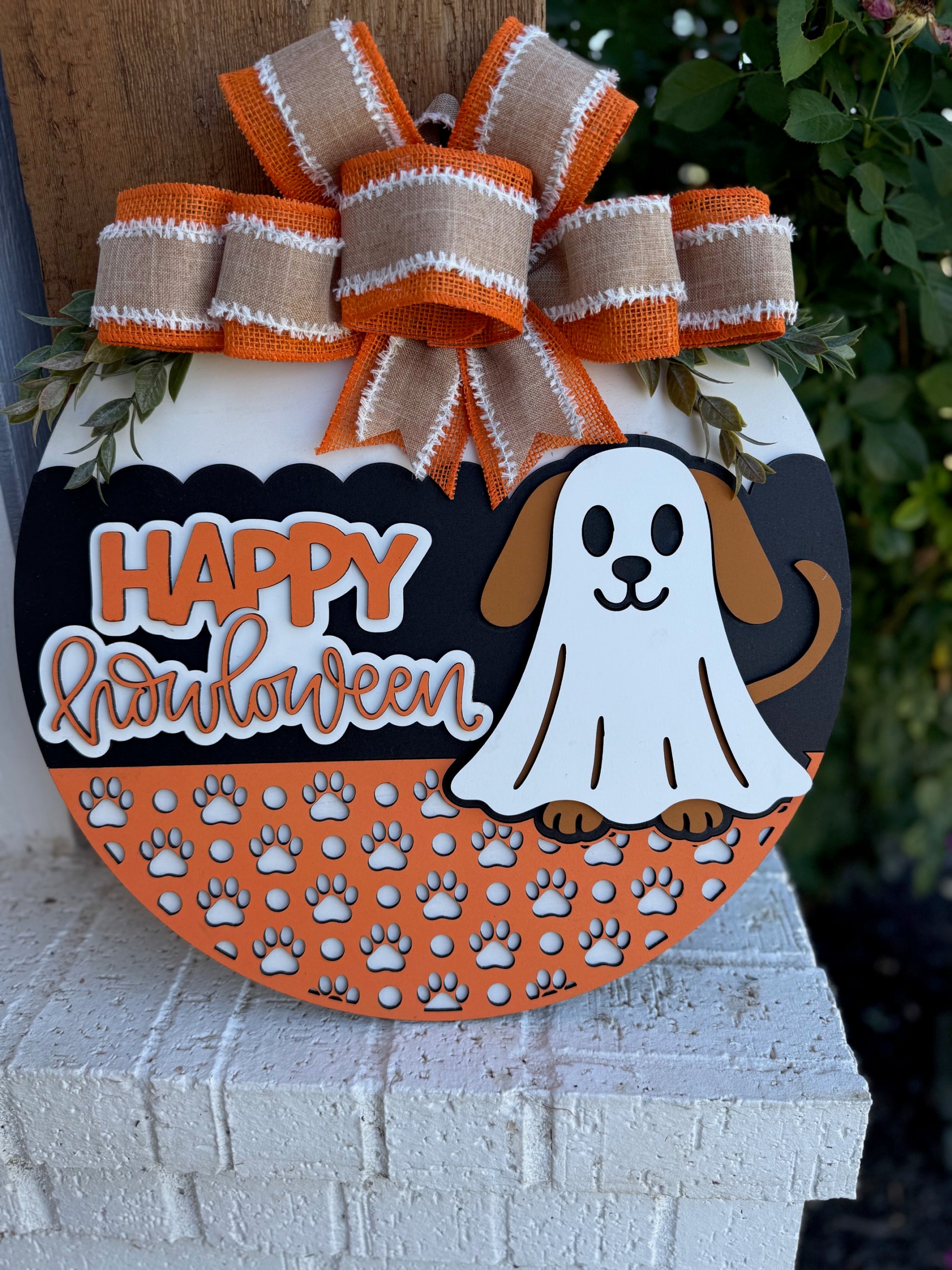 A Halloween-themed round sign with a cartoon dog in a ghost costume, orange bow on top, and the words Happy Howloween above a pattern of white paw prints. The sign rests on a white brick surface with greenery behind it.
