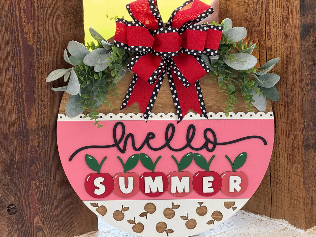 A round, decorative wooden sign hangs on a door. It features greenery, a red and black polka-dot bow, the word “hello” in cursive, and “SUMMER” spelled with white letters on red apple shapes. The lower section has a pattern of small apples.