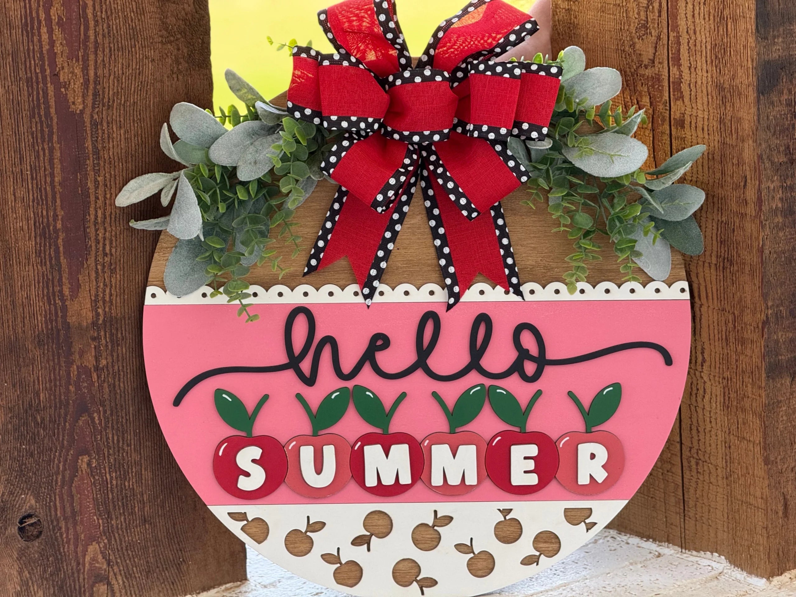 A round, decorative wooden sign hangs on a door. It features greenery, a red and black polka-dot bow, the word “hello” in cursive, and “SUMMER” spelled with white letters on red apple shapes. The lower section has a pattern of small apples.
