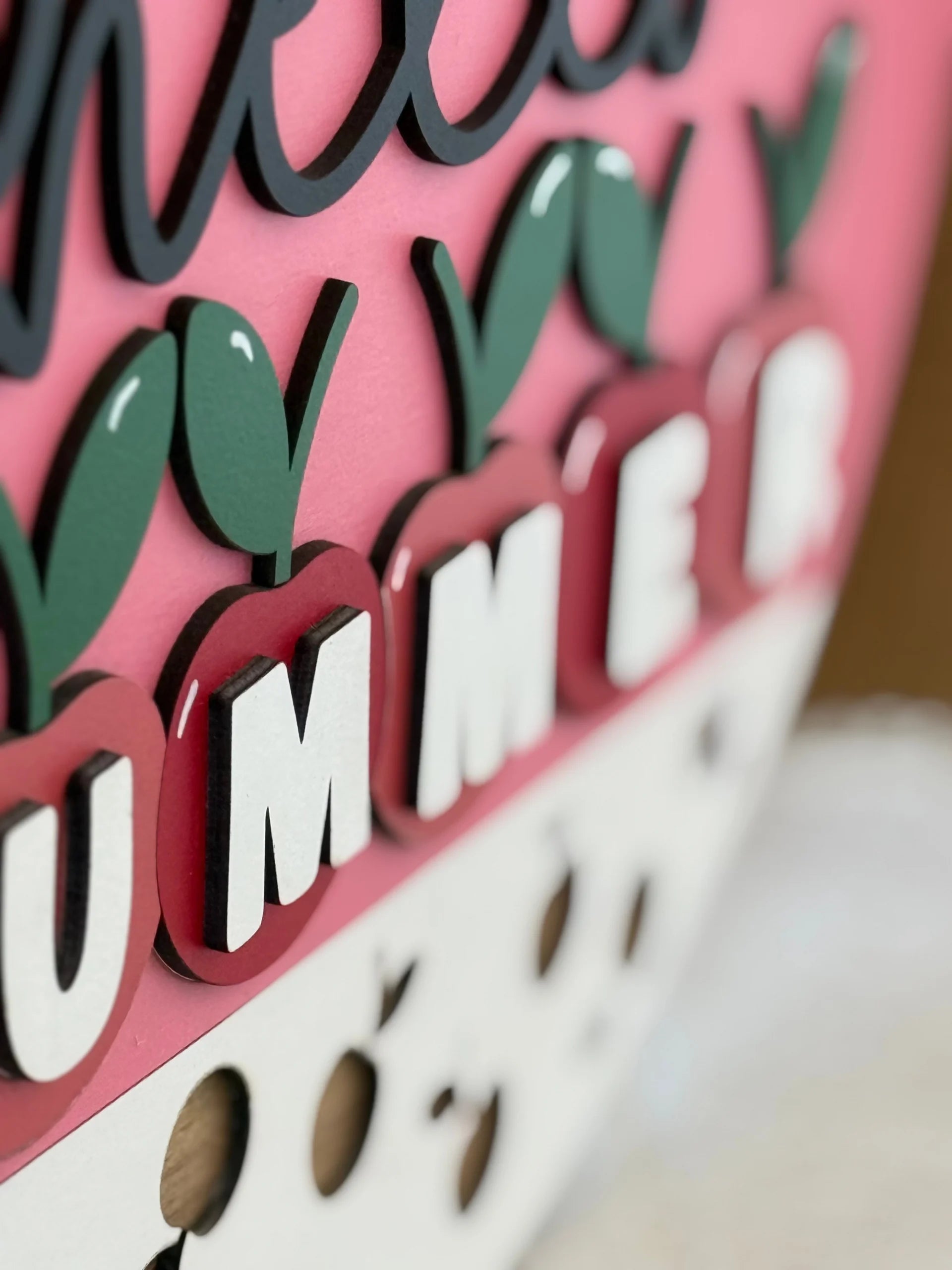 Close-up of a colorful, layered sign with a pink background. Black script and bold white block letters spell out SUMMER, with each letter mounted on red cherries amid green leaves. The bottom has a white section with small, round cutouts.