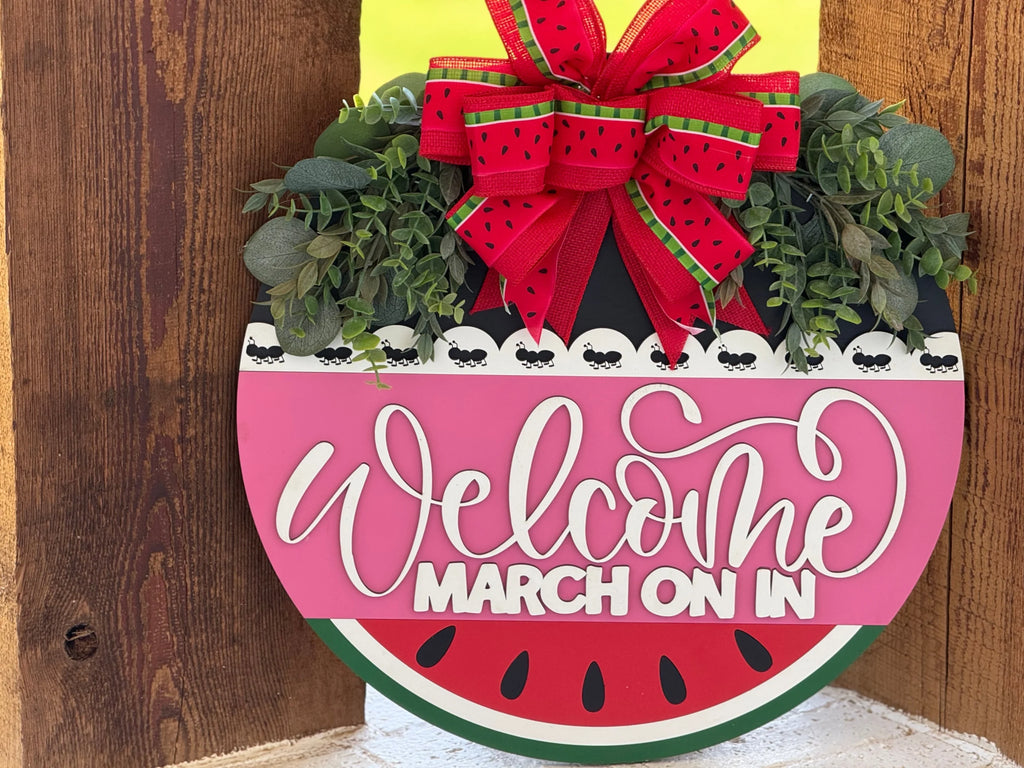 A round sign resembling a watermelon slice hangs on wood. It features a red, green, and white design, leafy greenery, and a large red bow with watermelon patterns on top. The text reads Welcome MARCH ON IN in white and bold red letters.