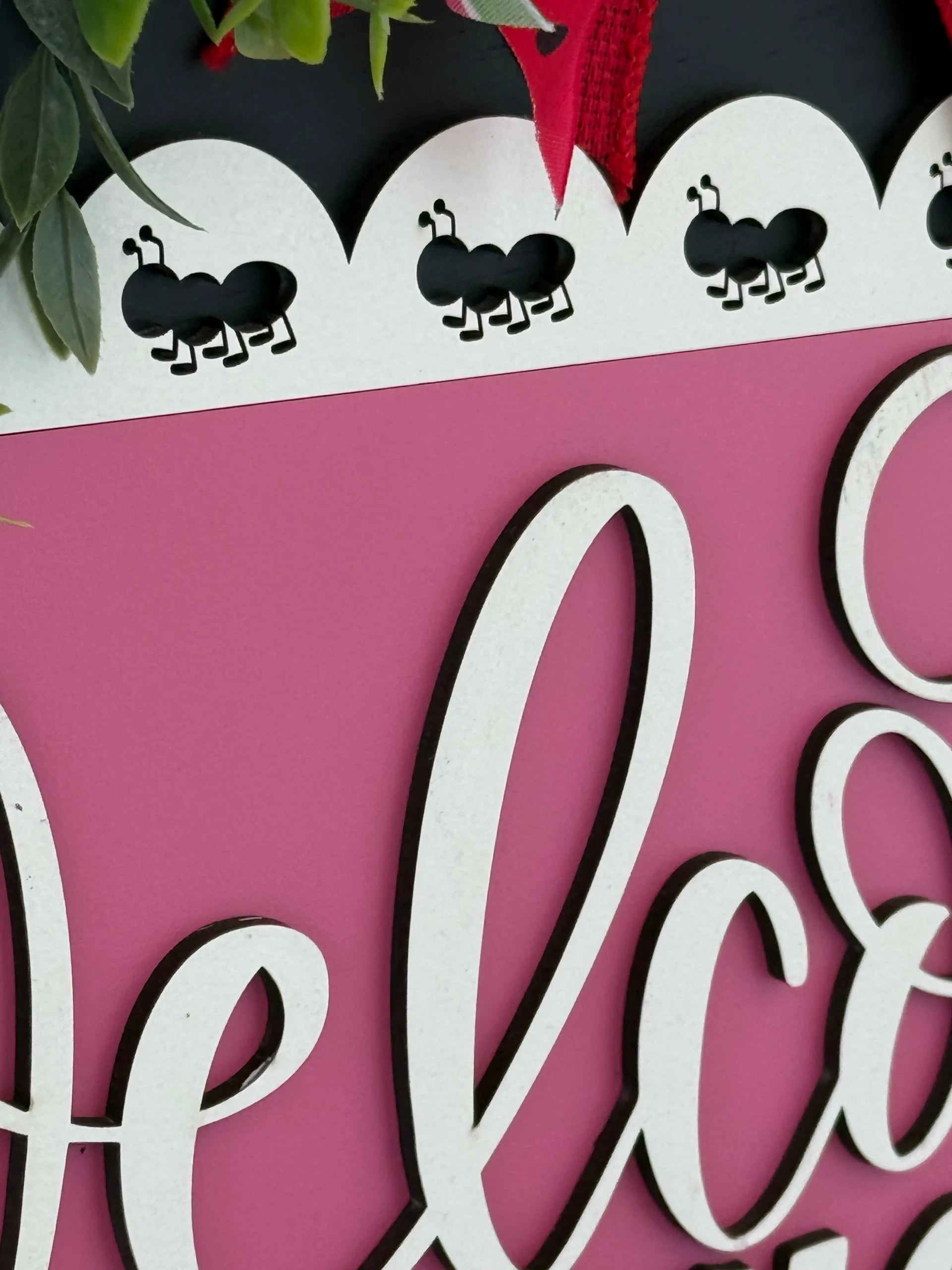 A close-up of a decorative sign with a scalloped white border featuring cutout ant designs. The sign is pink with large, cursive, white text, partially visible. Green leaves and a bit of red ribbon are seen at the top edge.