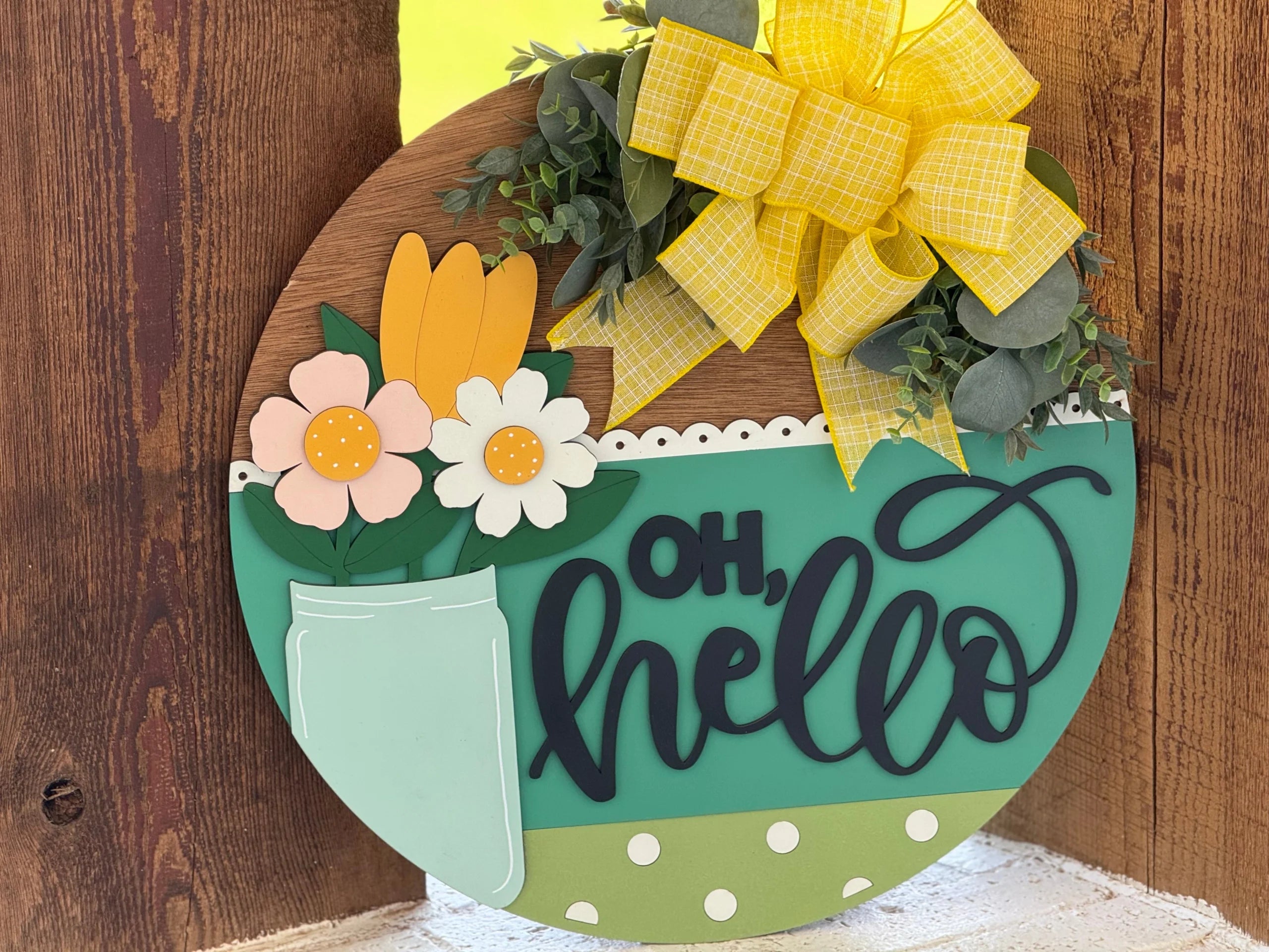 A round wooden sign decorated with a blue-green section reading “oh, hello” in black script. It features a painted mason jar with pastel flowers, green leaves, and a large yellow plaid bow at the top, leaning against a rustic wooden surface.