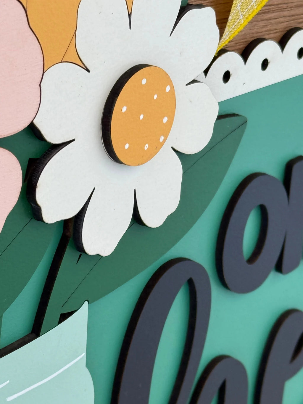 A close-up of a decorative wooden sign features a white daisy with a yellow center and small white dots, green leaves, and part of the word on in bold black letters on a green background. Part of a pink flower and lace-like trim are also visible.