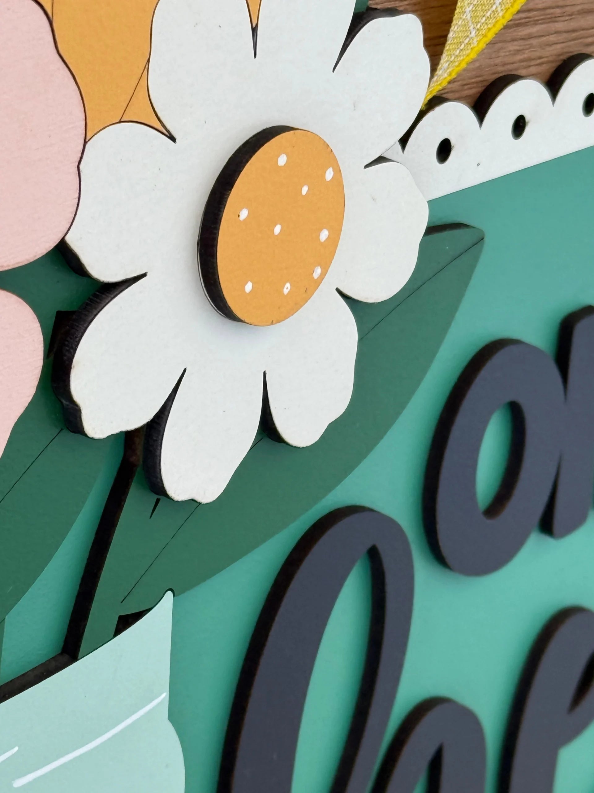 A close-up of a decorative wooden sign features a white daisy with a yellow center and small white dots, green leaves, and part of the word on in bold black letters on a green background. Part of a pink flower and lace-like trim are also visible.