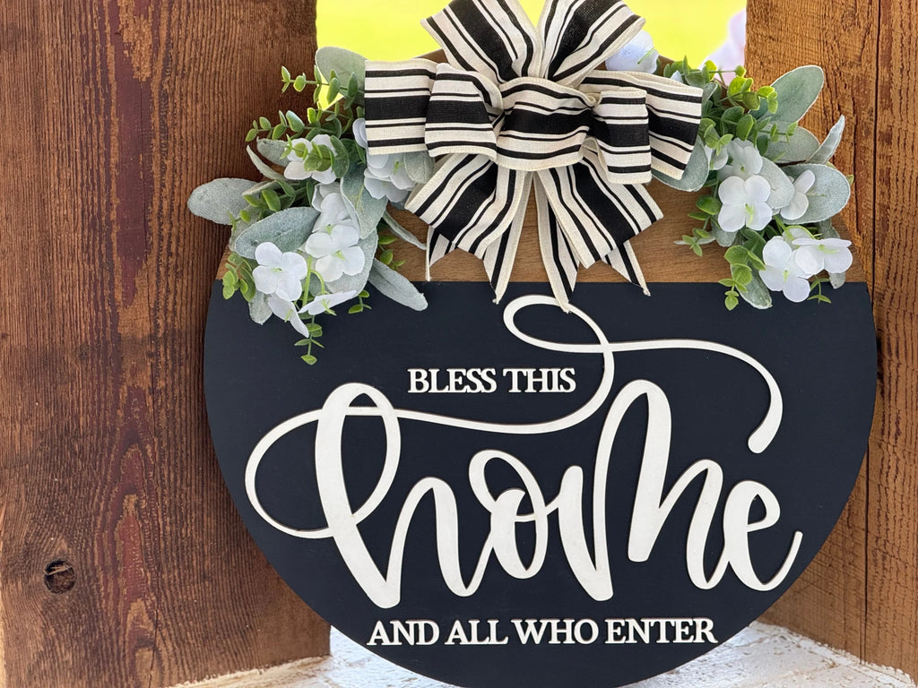 A round black sign with “Bless this home and all who enter” in white script sits on a rustic wooden background. The sign is decorated with a black-and-white striped bow, faux green leaves, and white flowers at the top.