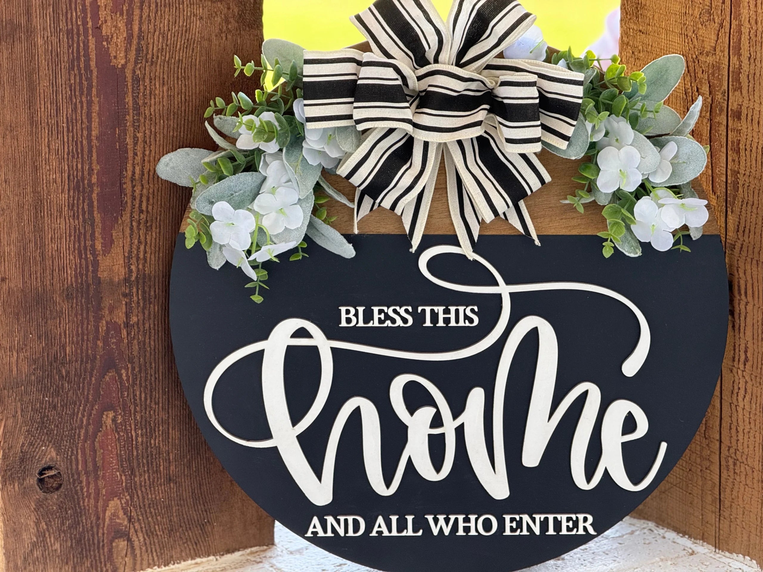 A round black sign with “Bless this home and all who enter” in white script sits on a rustic wooden background. The sign is decorated with a black-and-white striped bow, faux green leaves, and white flowers at the top.
