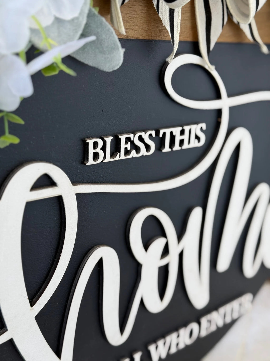 A decorative black sign with raised white script reads BLESS THIS at the top, followed by large, partially visible cursive text. Faux white flowers and green leaves adorn the upper corner, and wooden elements appear at the top edge.