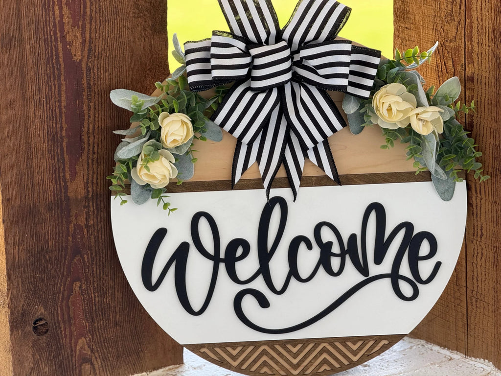 A wooden “Welcome” sign hangs on a wall. It’s half-round, decorated with yellow roses, green foliage, and topped with a large black-and-white striped bow. The background includes brown wood planks. The word “Welcome” is written in curly black script.