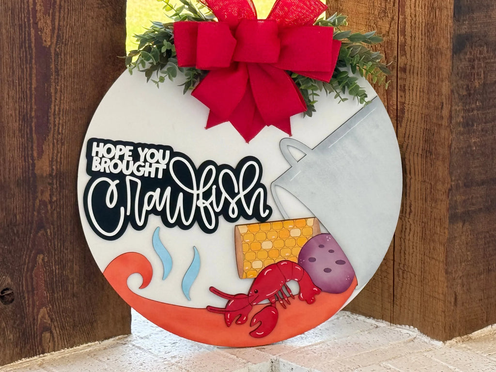 A round door hanger with a red bow, greenery, and the text Hope you brought crawfish. It features a pot, corn, potatoes, steam curls, and a red crawfish on an orange swirl, set against a wooden background.