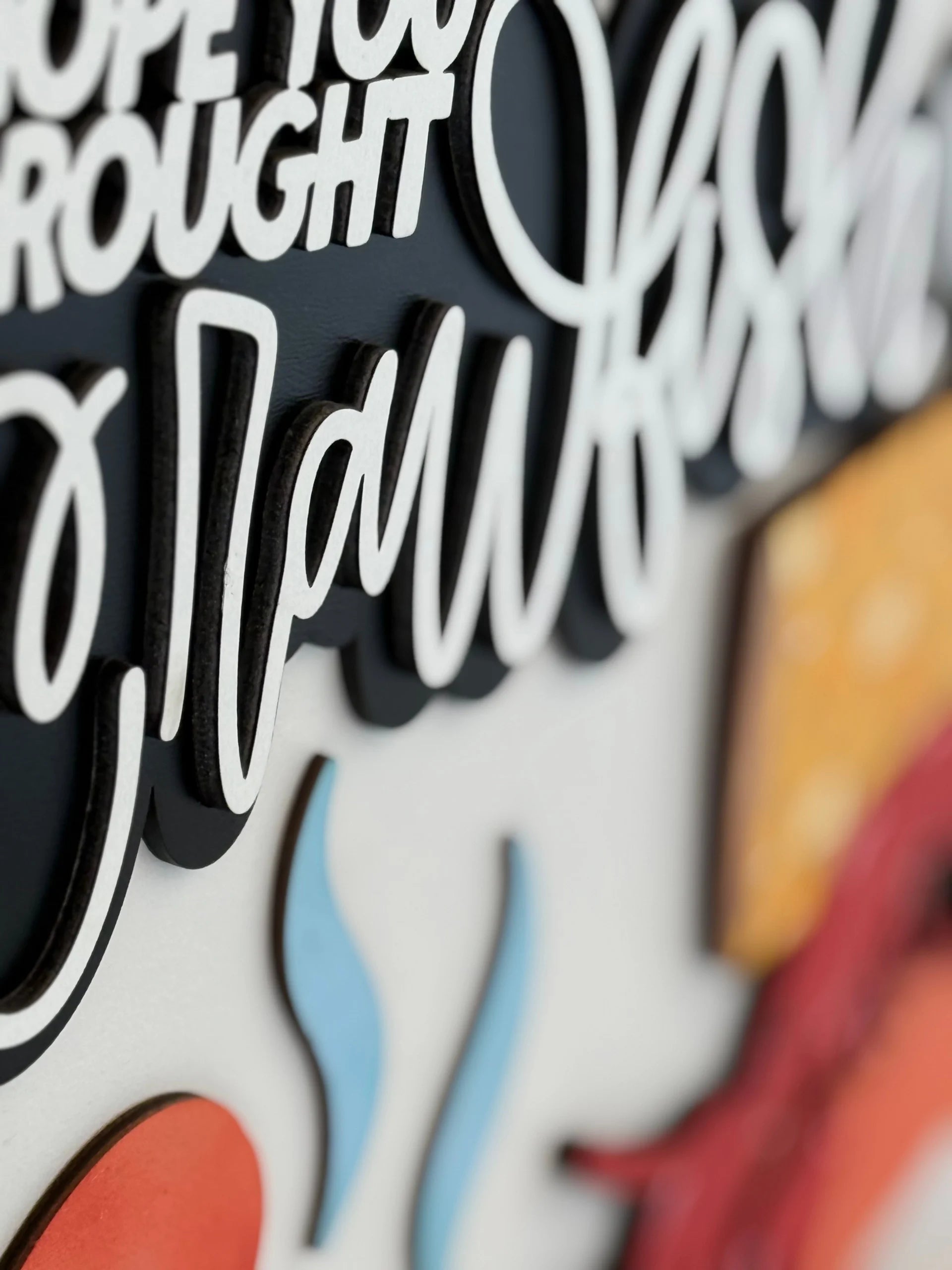 Close-up of a layered sign with bold, raised, black-and-white cursive text that says crawfish. Decorative elements include a red crawfish claw, yellow corn, and blue wavy lines, all made from cutout pieces for a 3D effect.