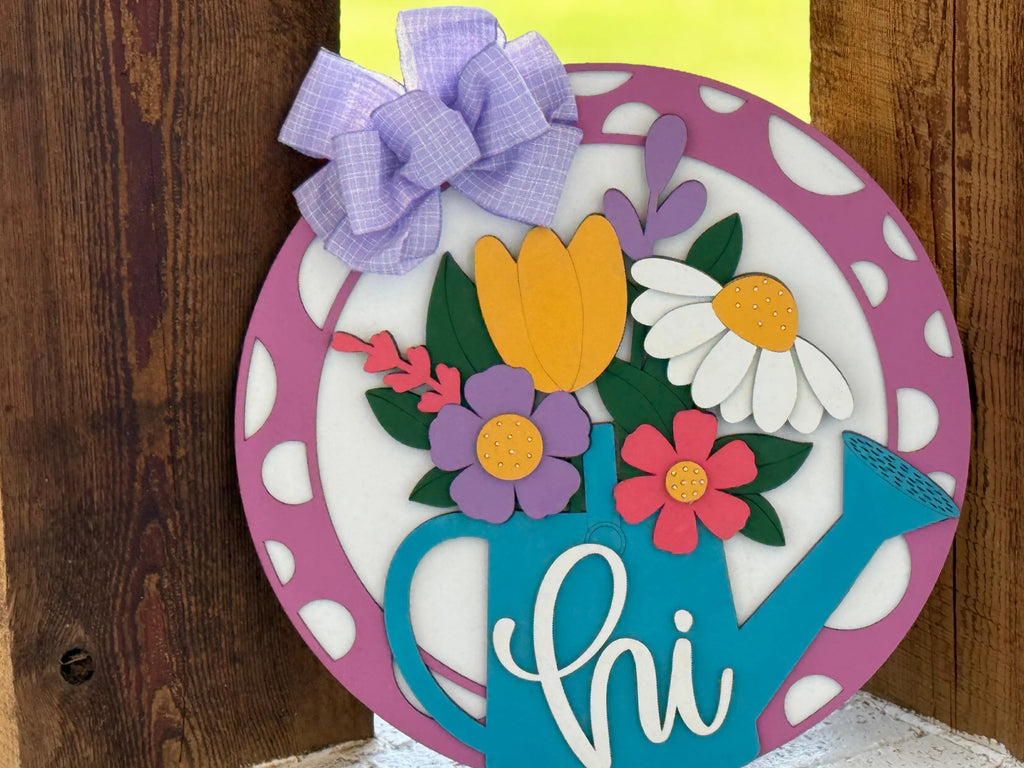 A round, decorative sign features a blue watering can with hi written on it, filled with colorful flowers. The sign has a purple and white polka dot border and is topped with a lavender plaid bow, leaning against rustic wooden planks.