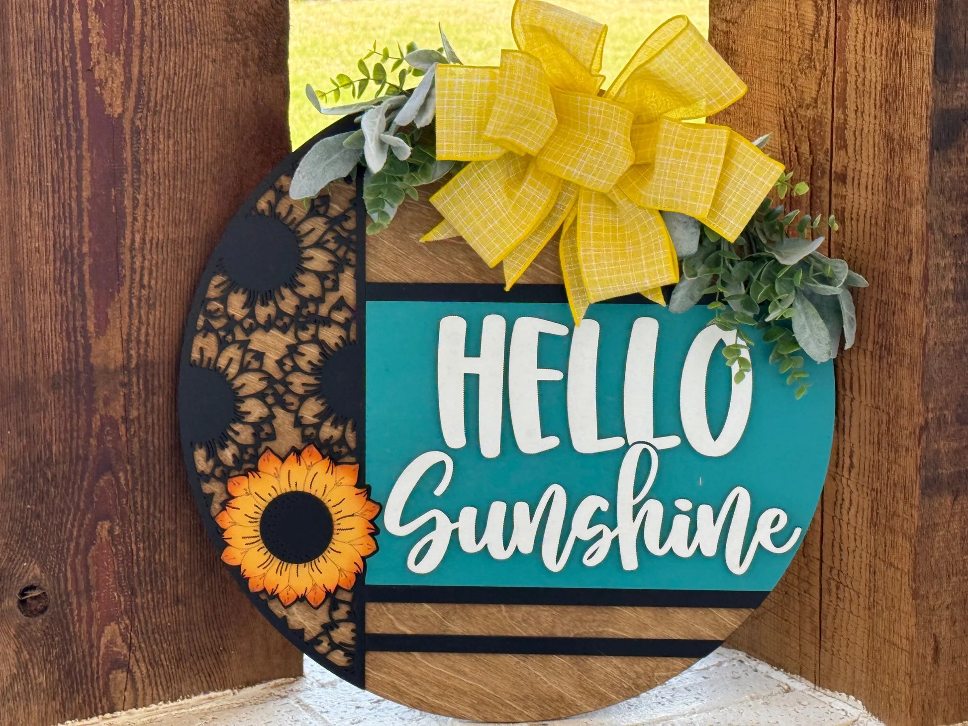 A round wooden sign features the words “HELLO Sunshine” in bold white letters on a teal background. The sign has sunflower designs on the left, a bright yellow plaid bow, and green foliage at the top, and leans against a rustic wooden backdrop.