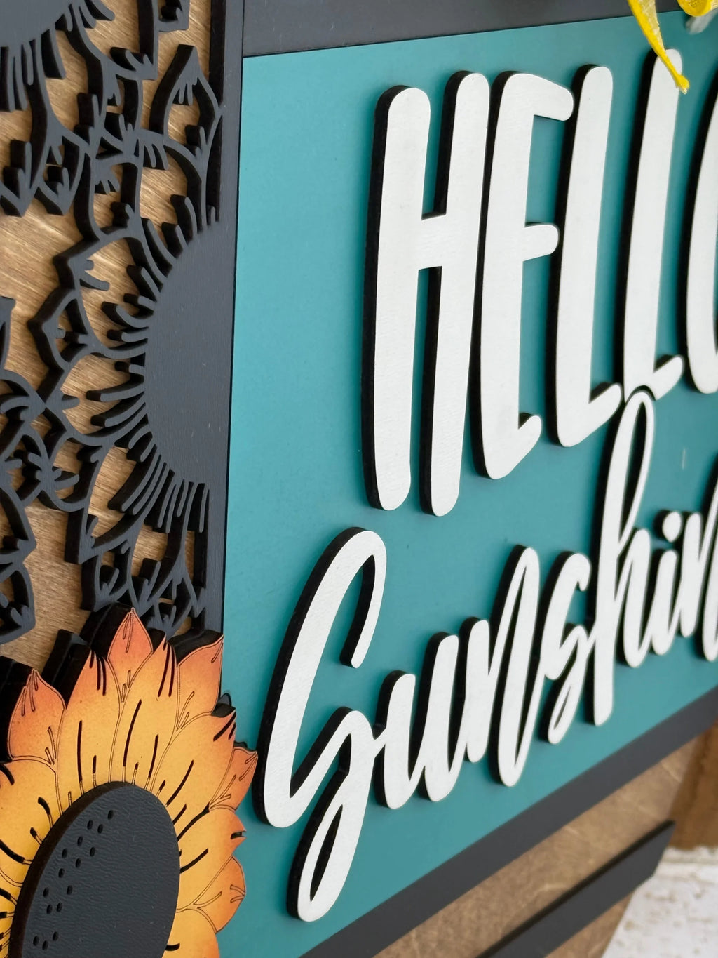 A close-up of a decorative sign with bold, white raised text reading “Hello Sunshine” on a teal background. The sign features a detailed black floral pattern on the left and a painted sunflower with orange petals in the bottom left corner.