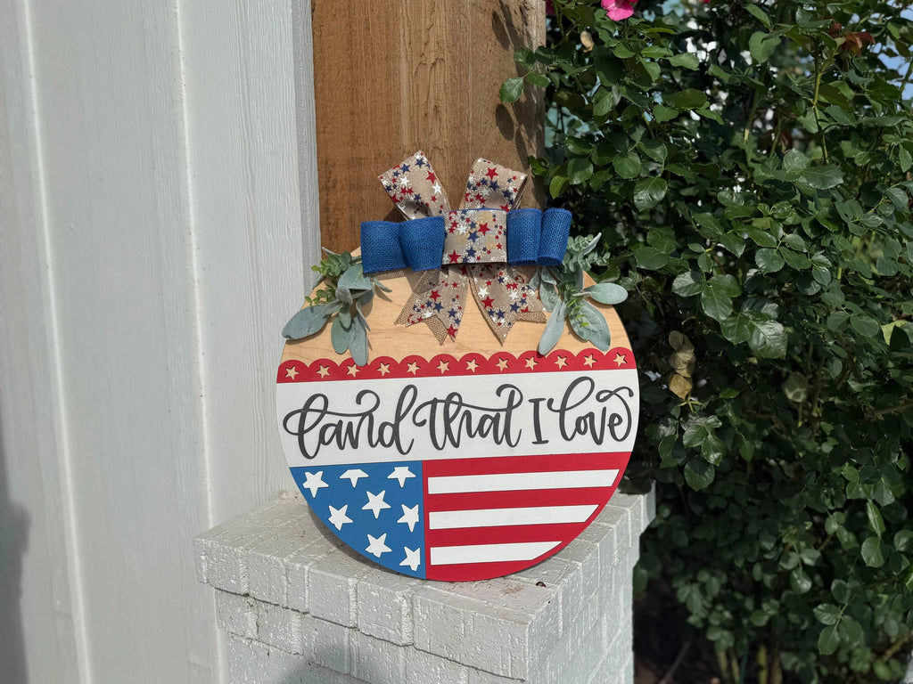 A round wooden sign with stars, stripes, and the words land that I love leans against an outdoor post. It is decorated with a bow and greenery, with roses and leaves in the background.