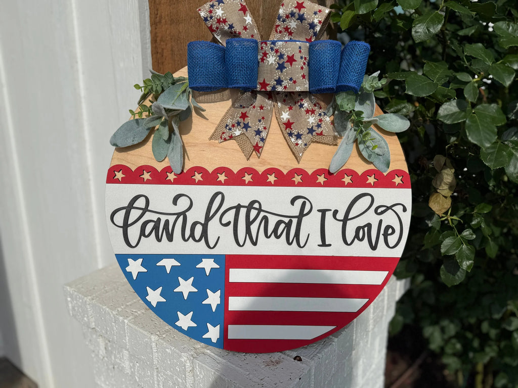 A patriotic door sign shaped like a heart, decorated with stars, stripes, greenery, and a festive bow. It reads “Land that I love” in cursive. The background includes greenery and part of a white wall.