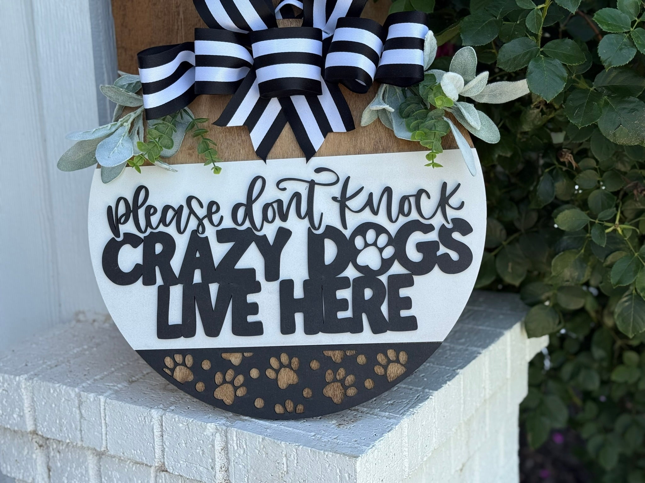 A round sign with a striped bow reads “Please don’t knock, crazy dogs live here” with paw prints and leaves, hanging on a white brick porch next to green foliage.