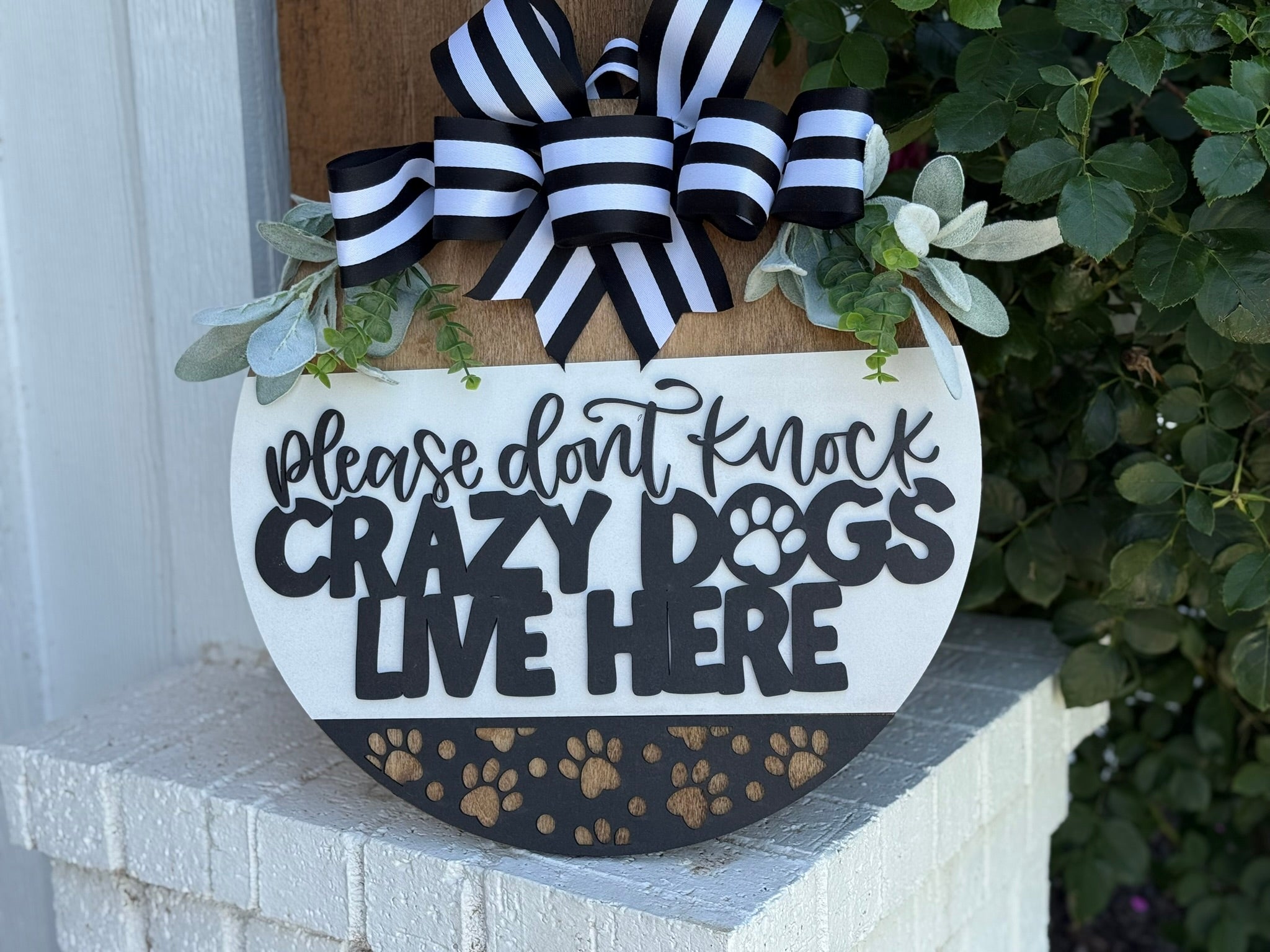 A round wooden sign with black and white striped bows, greenery, and paw prints reads: “Please don’t knock. Crazy dogs live here.” The sign hangs on a white brick wall near green leafy plants.