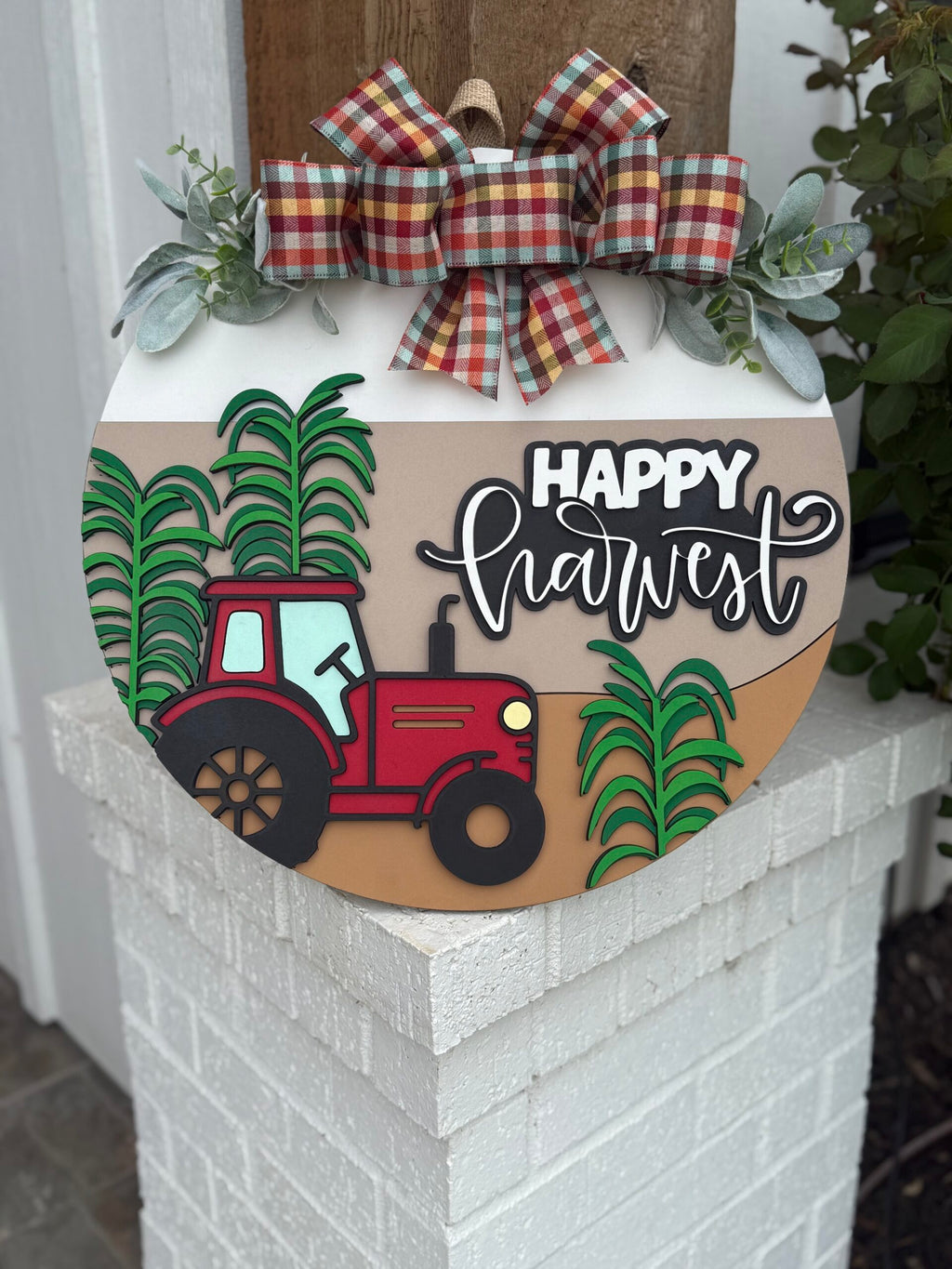 A round sign decorated with a red tractor, green corn stalks, and the words Happy Harvest in cursive. It features a plaid bow and greenery at the top, and is displayed on a white brick post.