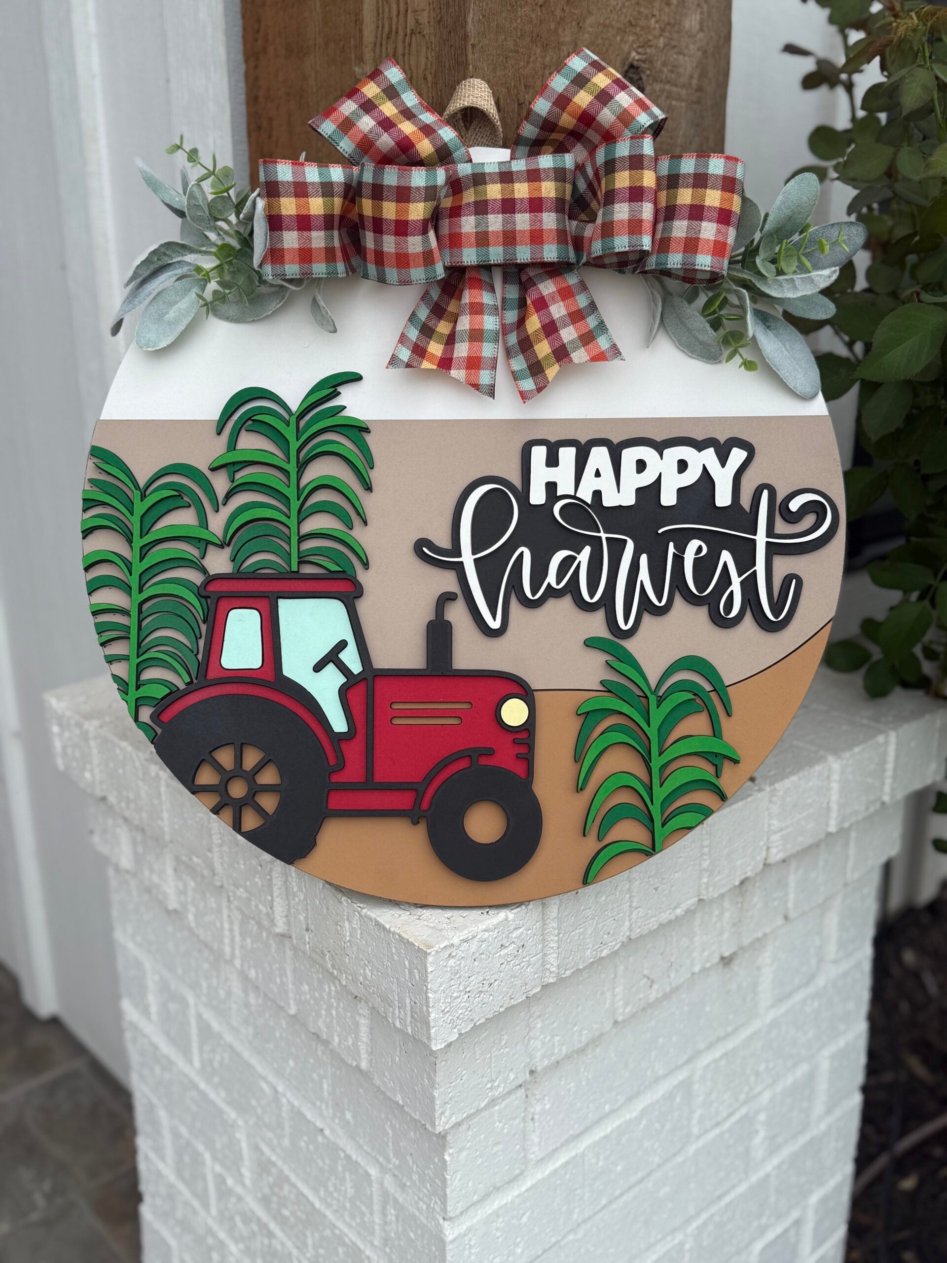 A round sign decorated with a red tractor, green corn stalks, and the words Happy Harvest in cursive. It features a plaid bow and greenery at the top, and is displayed on a white brick post.