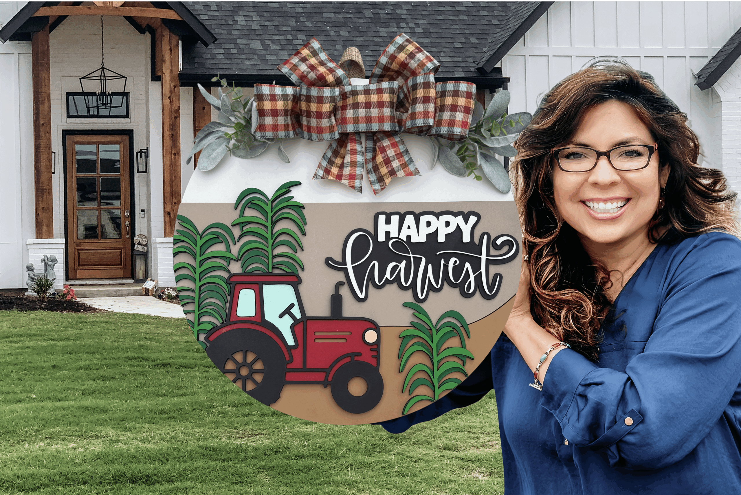 A smiling woman in glasses and a blue top holds a round sign decorated with a red tractor, green crops, a plaid bow, and the words Happy Harvest in front of a modern farmhouse.