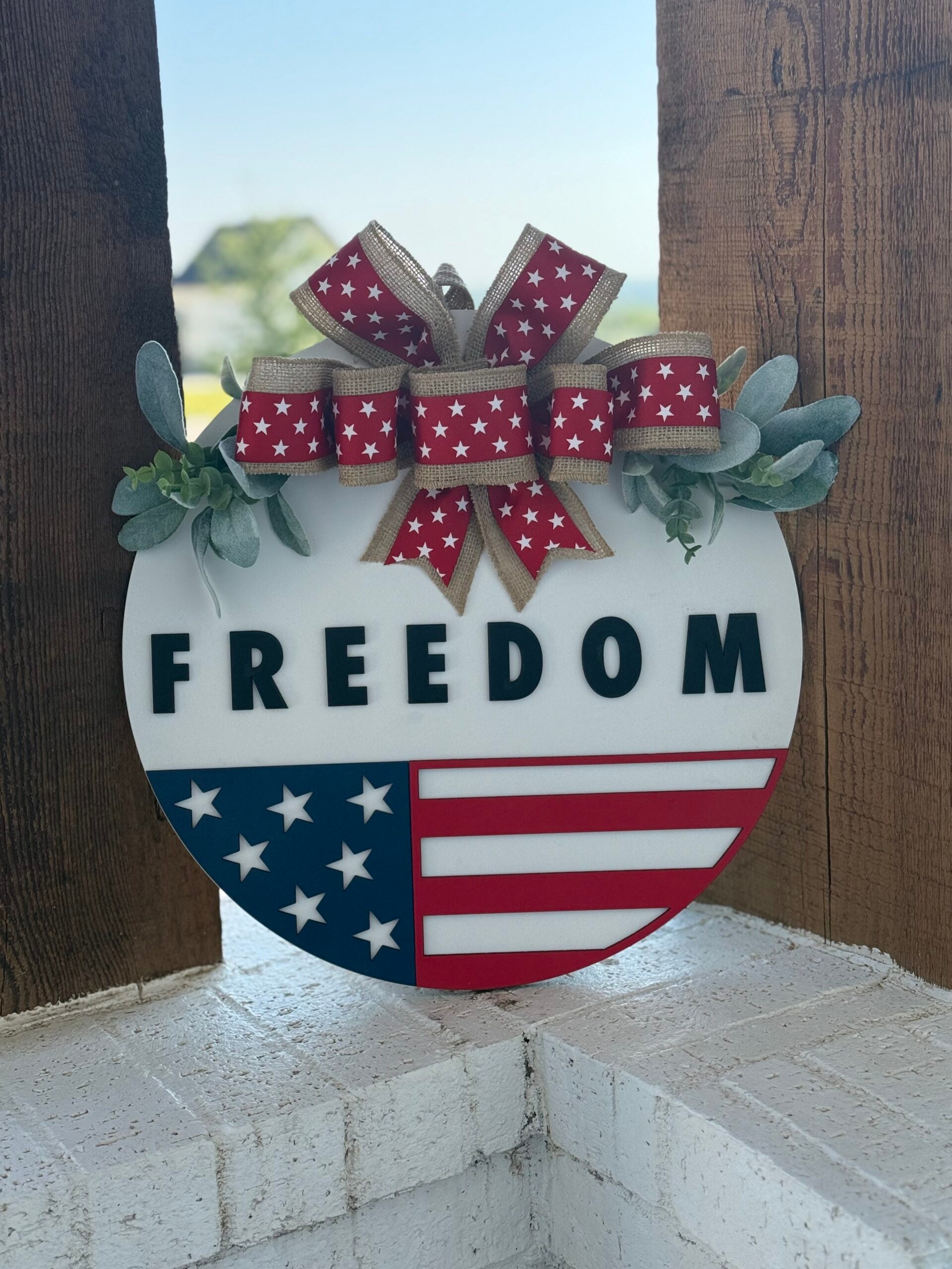 A round sign with FREEDOM in bold letters, decorated with a red, white, and blue American flag motif, green foliage, and a star-patterned bow, hanging on a wooden post.