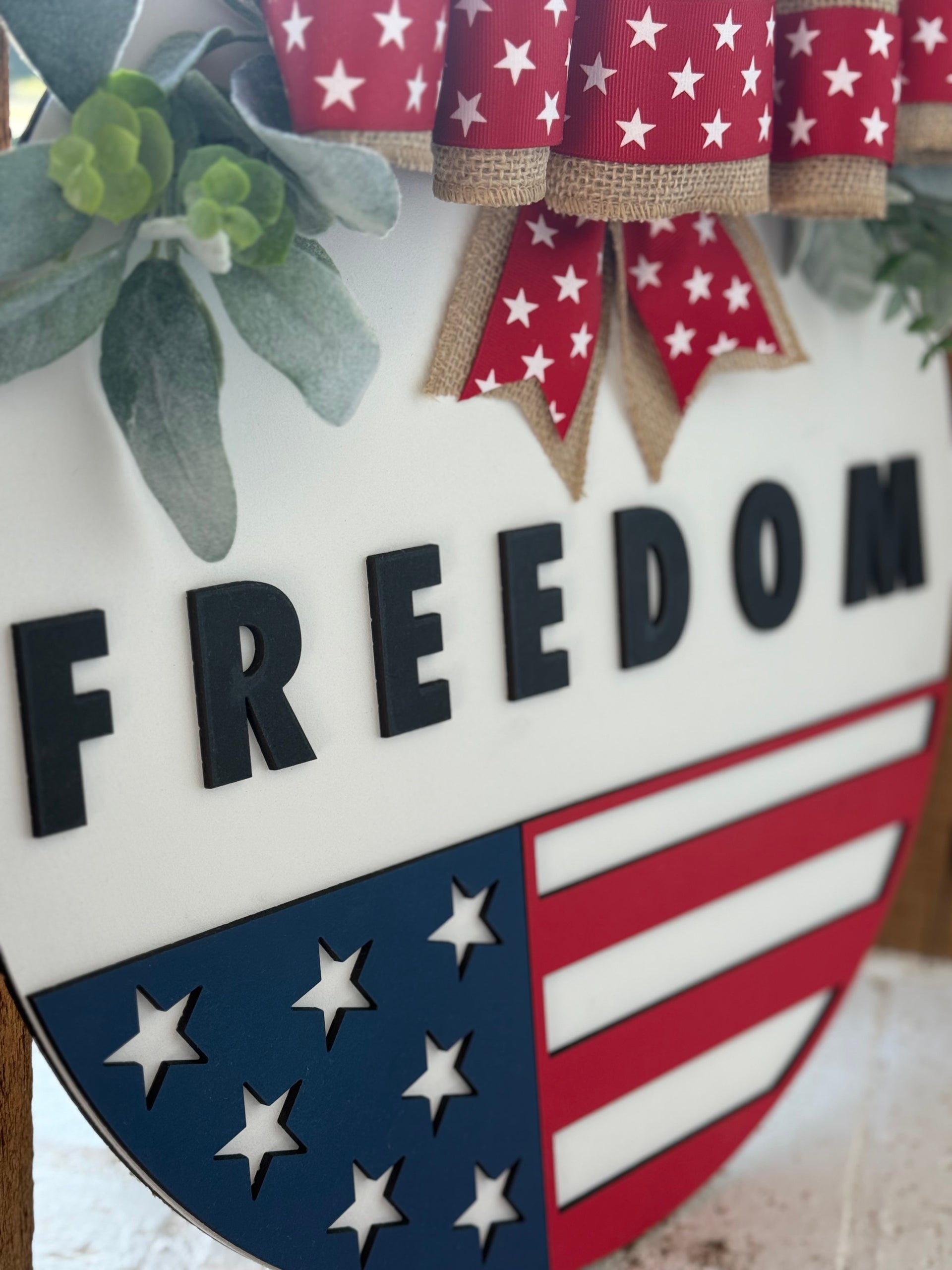 A decorative sign with the word FREEDOM in bold black letters, red and white ribbon with white stars, green leaves, and a shield featuring stars and stripes in blue, white, and red, resembling the American flag.