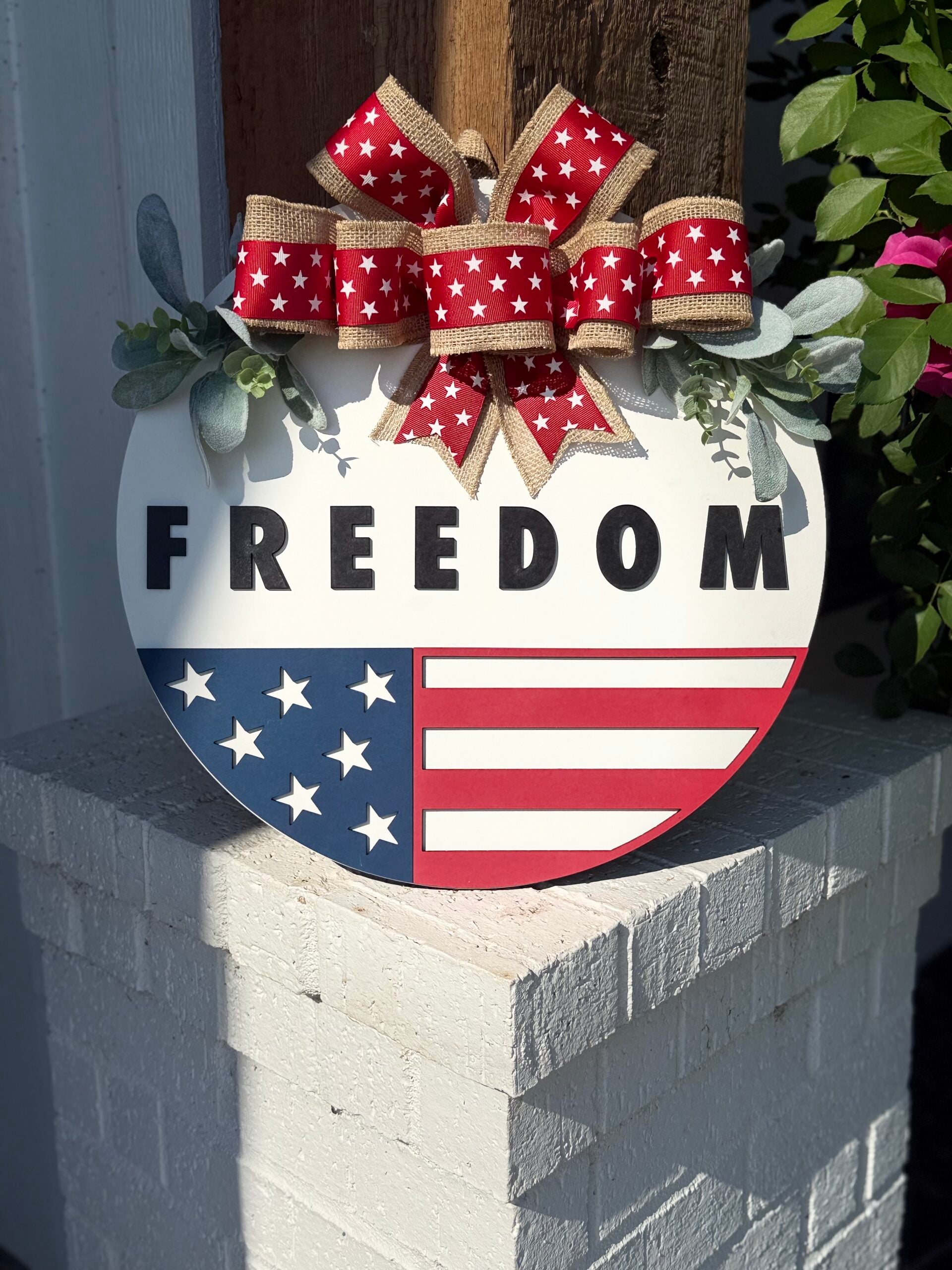 A round sign with the word FREEDOM above a partial American flag design, topped with a red, white, and burlap bow with white stars, is displayed on a white brick surface near some green plants.