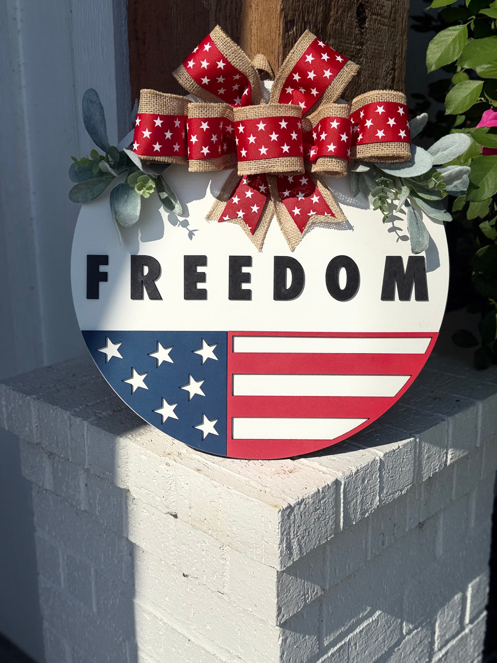 A round sign with FREEDOM in bold black letters above a stylized American flag, decorated with a red, white, and burlap bow with white stars, and greenery. The sign is displayed on a white brick ledge.