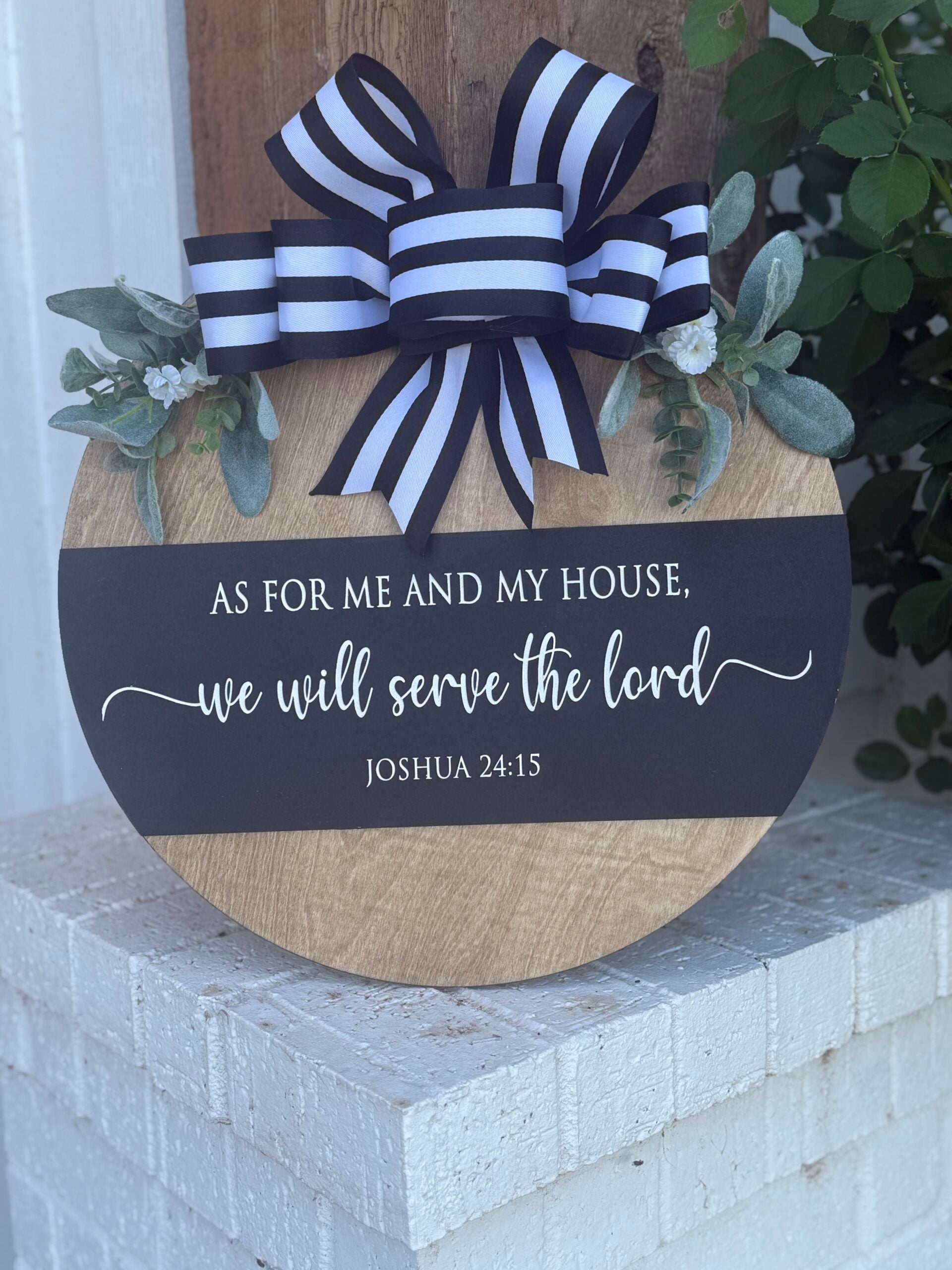 A round wooden sign with a black and white striped bow, greenery, and the text: As for me and my house, we will serve the Lord. Joshua 24:15, displayed on a white brick surface.