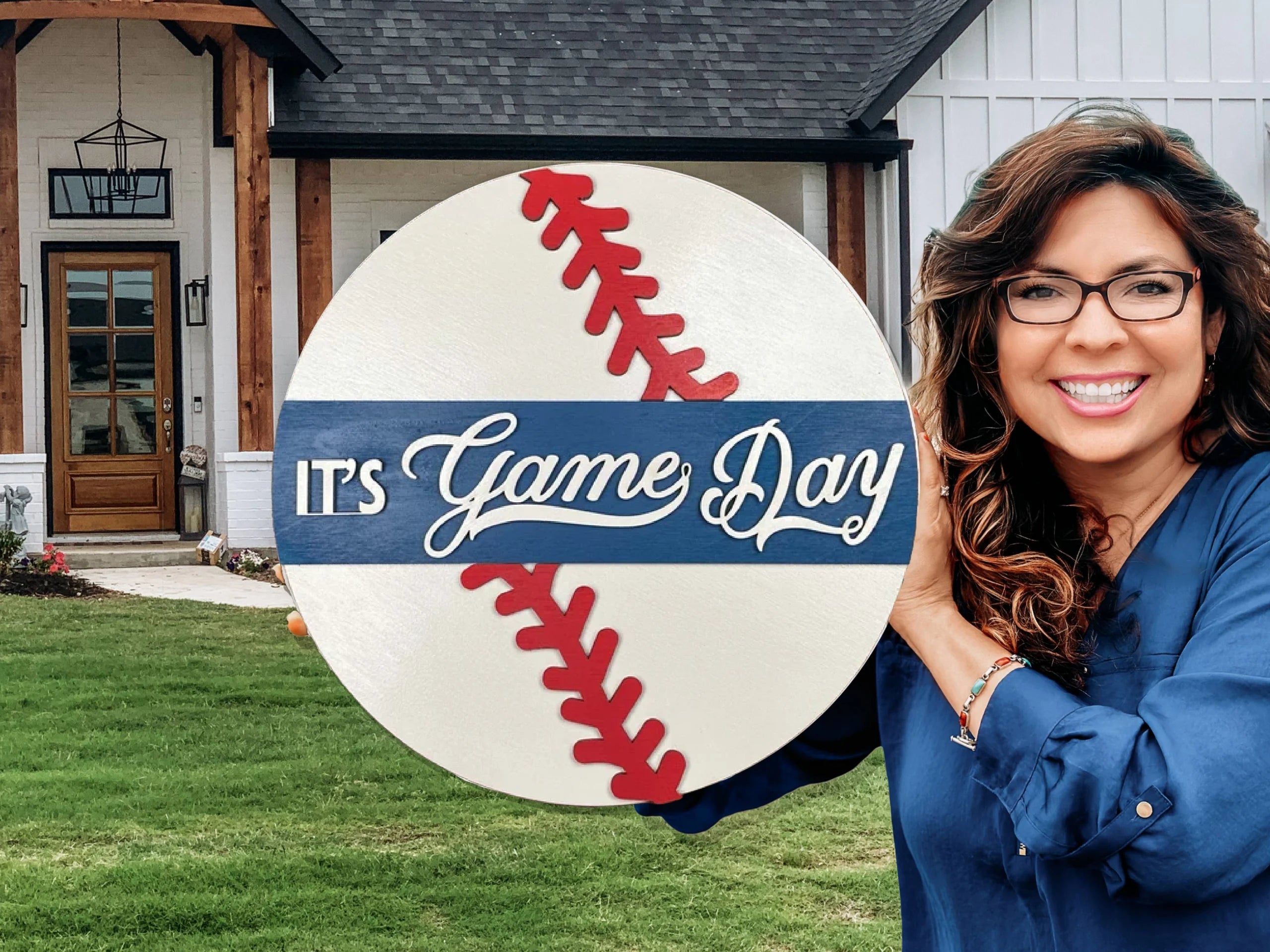 A smiling woman wearing glasses holds a round sign decorated like a baseball that reads “It’s Game Day” in front of a modern house with a green lawn.
