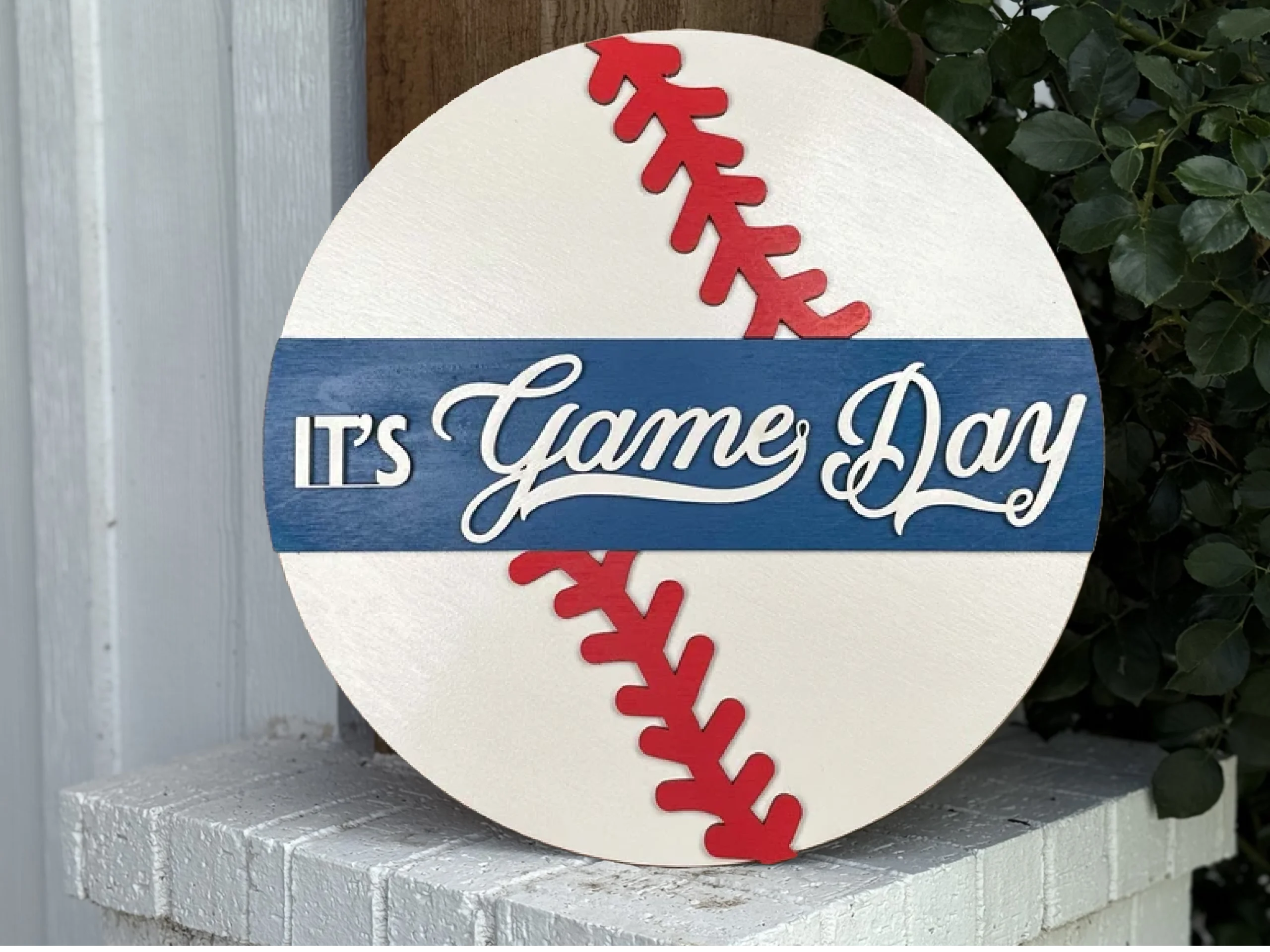 A round sign designed like a baseball with red stitching and a blue stripe across the middle. The sign reads ITS Game Day in white letters, and is propped against a white brick ledge near a plant.