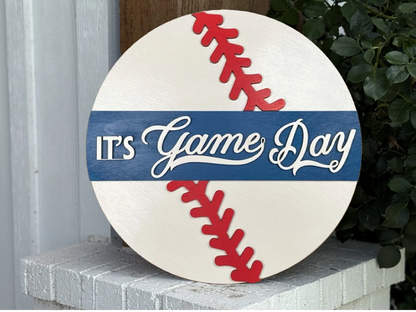 A round sign designed like a baseball with red stitching and a blue stripe across the middle. The sign reads ITS Game Day in white letters, and is propped against a white brick ledge near a plant.