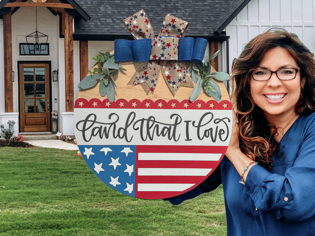 A smiling woman stands outside a house, holding a round wooden sign decorated with stars, stripes, greenery, and a bow. The sign reads Land that I love in cursive script.