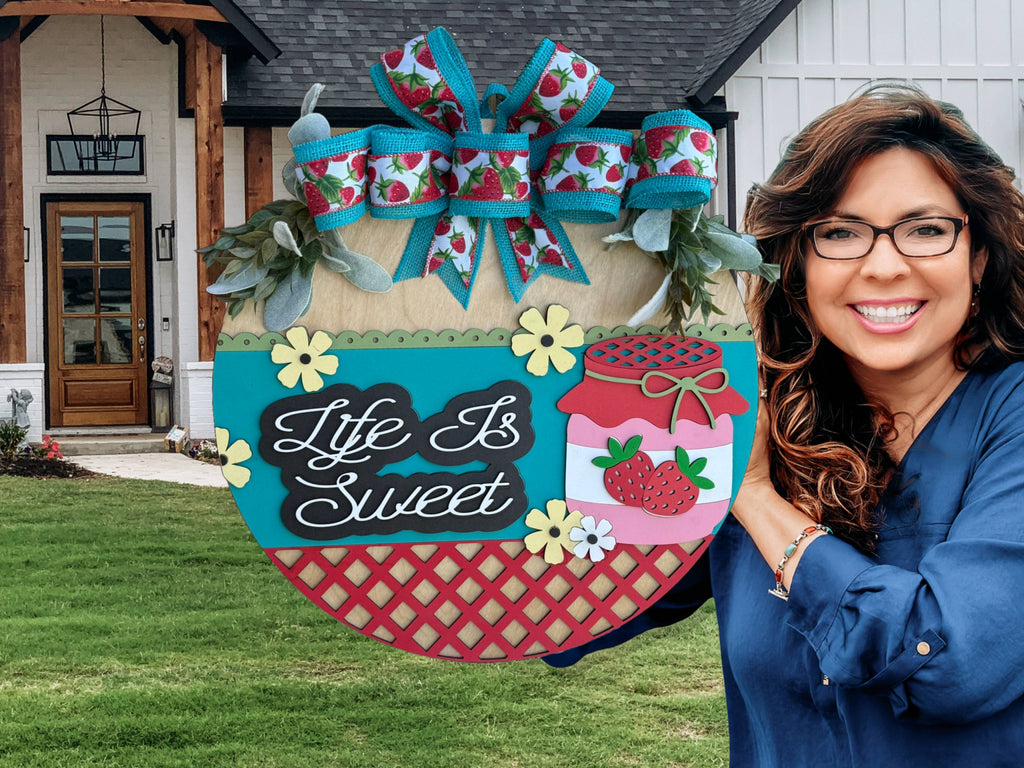 A smiling woman holds the Life Is Sweet Jam Jelly Door Sign, featuring bows, strawberries, and jars. Displayed outside a modern home, it offers a charming alternative to traditional door wreaths.