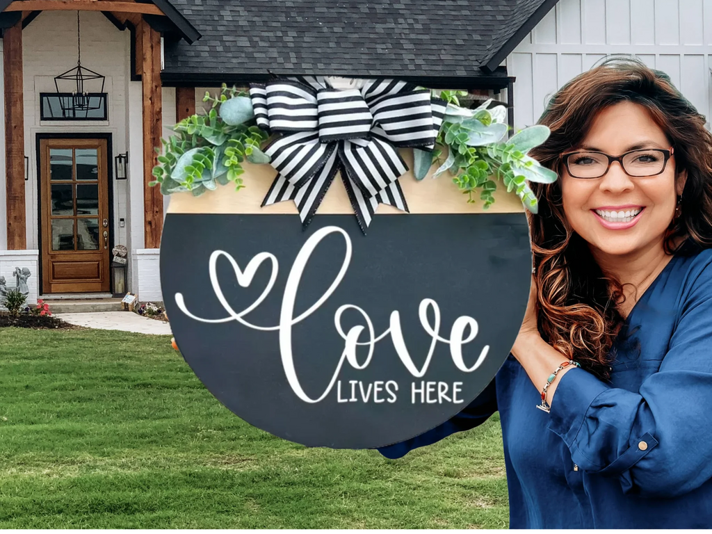 A smiling woman stands outside a modern house, holding a large round sign decorated with greenery, a striped bow, and the words “Love Lives Here” written in white script.