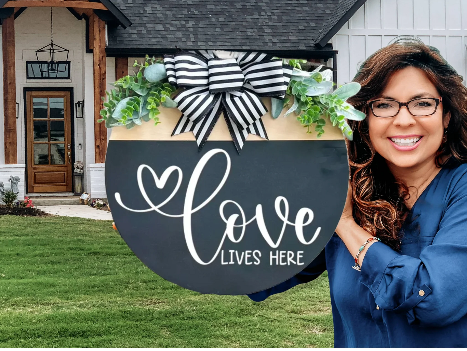 A smiling woman stands outside a modern house, holding a large round sign decorated with greenery, a striped bow, and the words “Love Lives Here” written in white script.